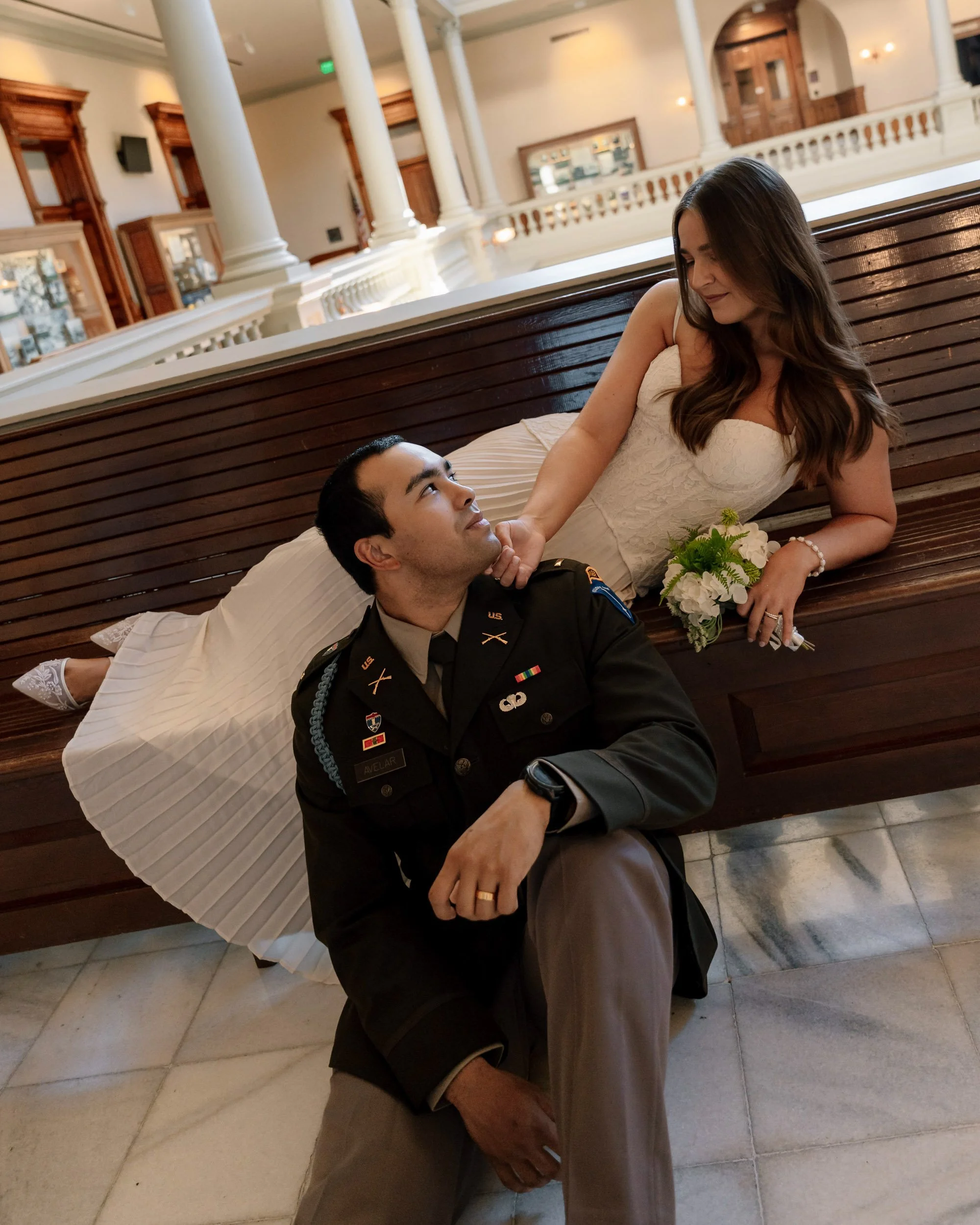 A woman in a white dress lying on a wooden bench, holding a small bouquet of flowers, looking at a man in military uniform who is sitting on the floor and gazing up at her, in a building with decorative columns and railings.