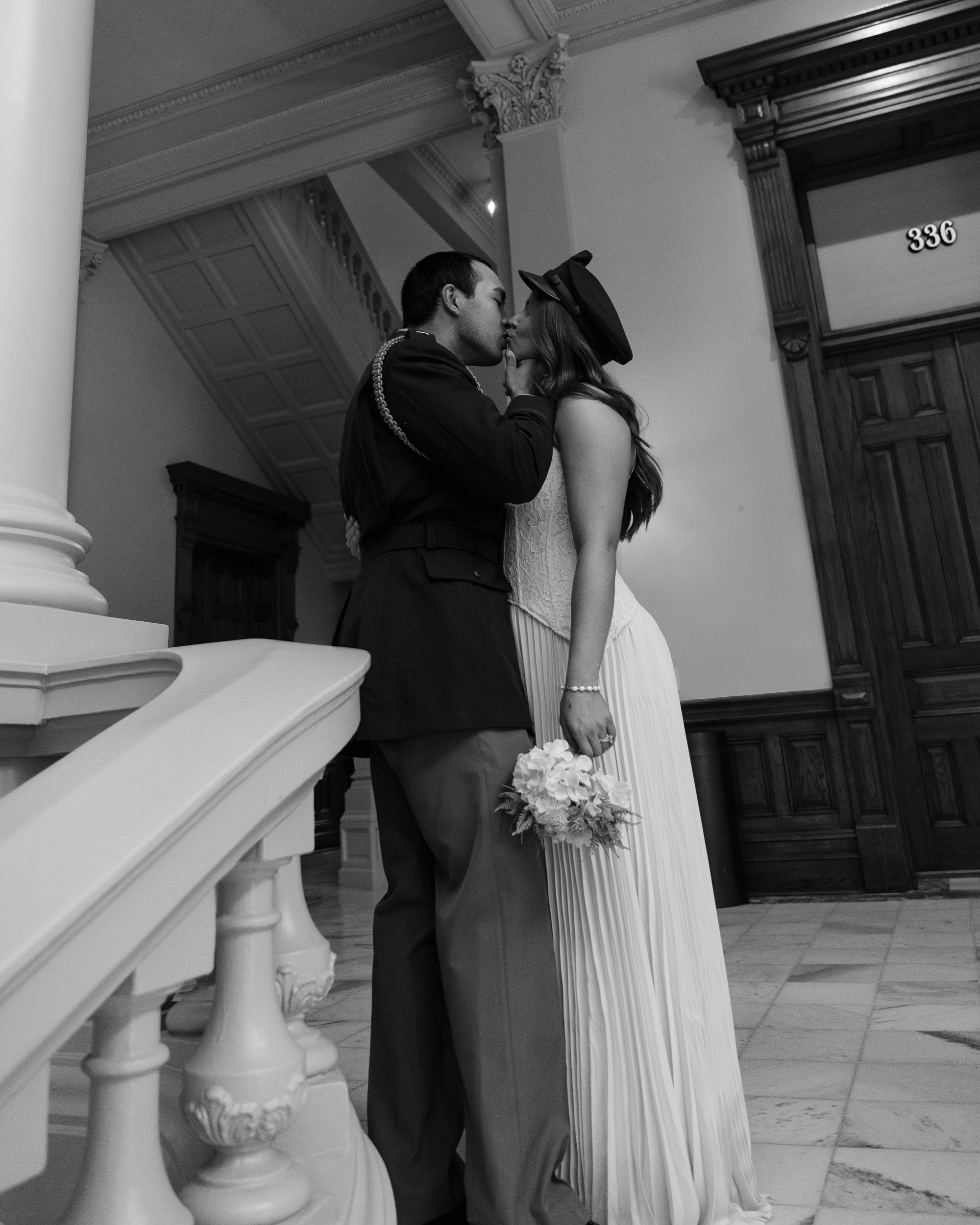 A black and white photo of a couple in wedding attire sharing a kiss inside a grand building with ornate architectural details.