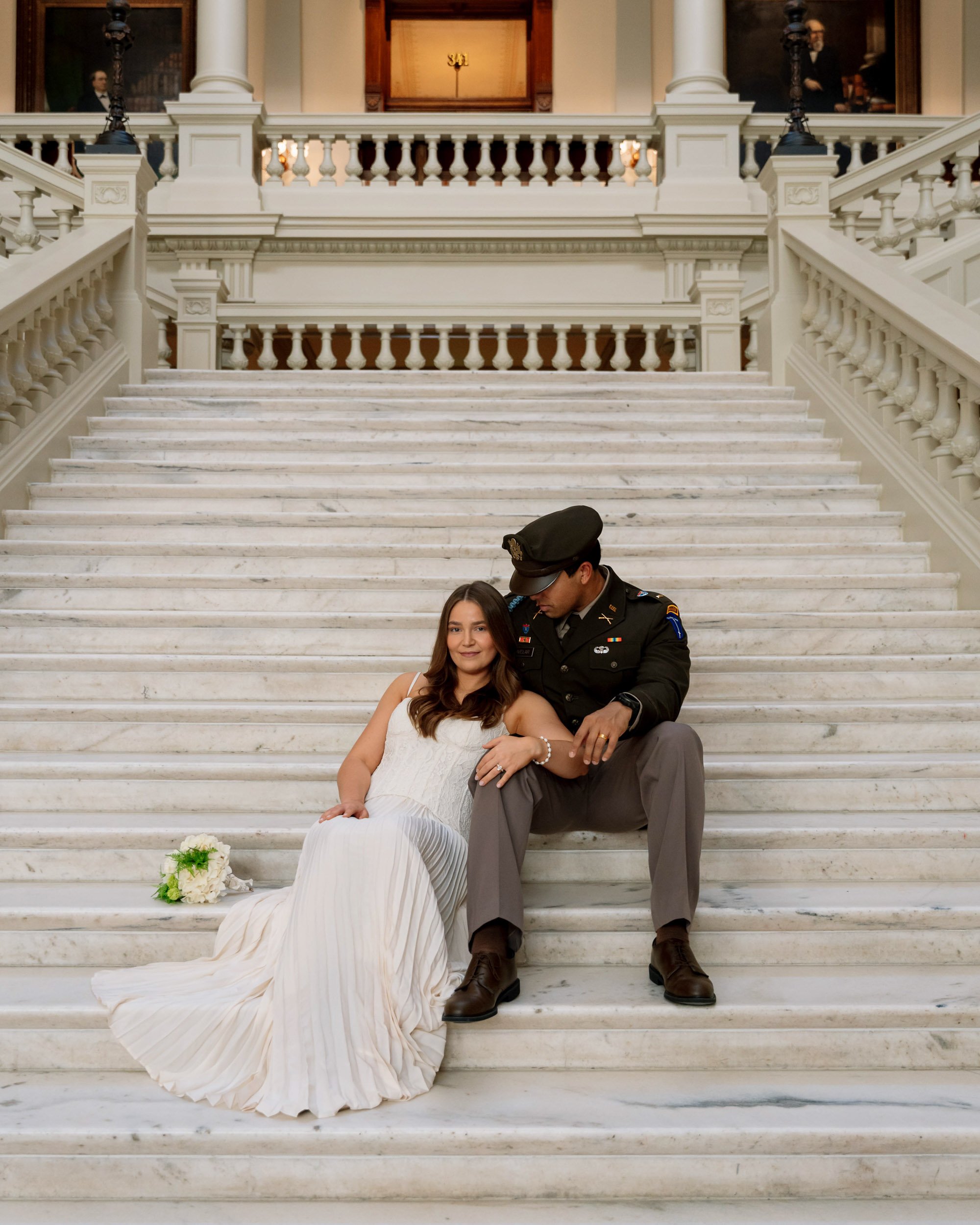 A woman in a white dress with a bouquet on the steps of a grand marble staircase, with a man in a military uniform sitting close, in an elegant building.