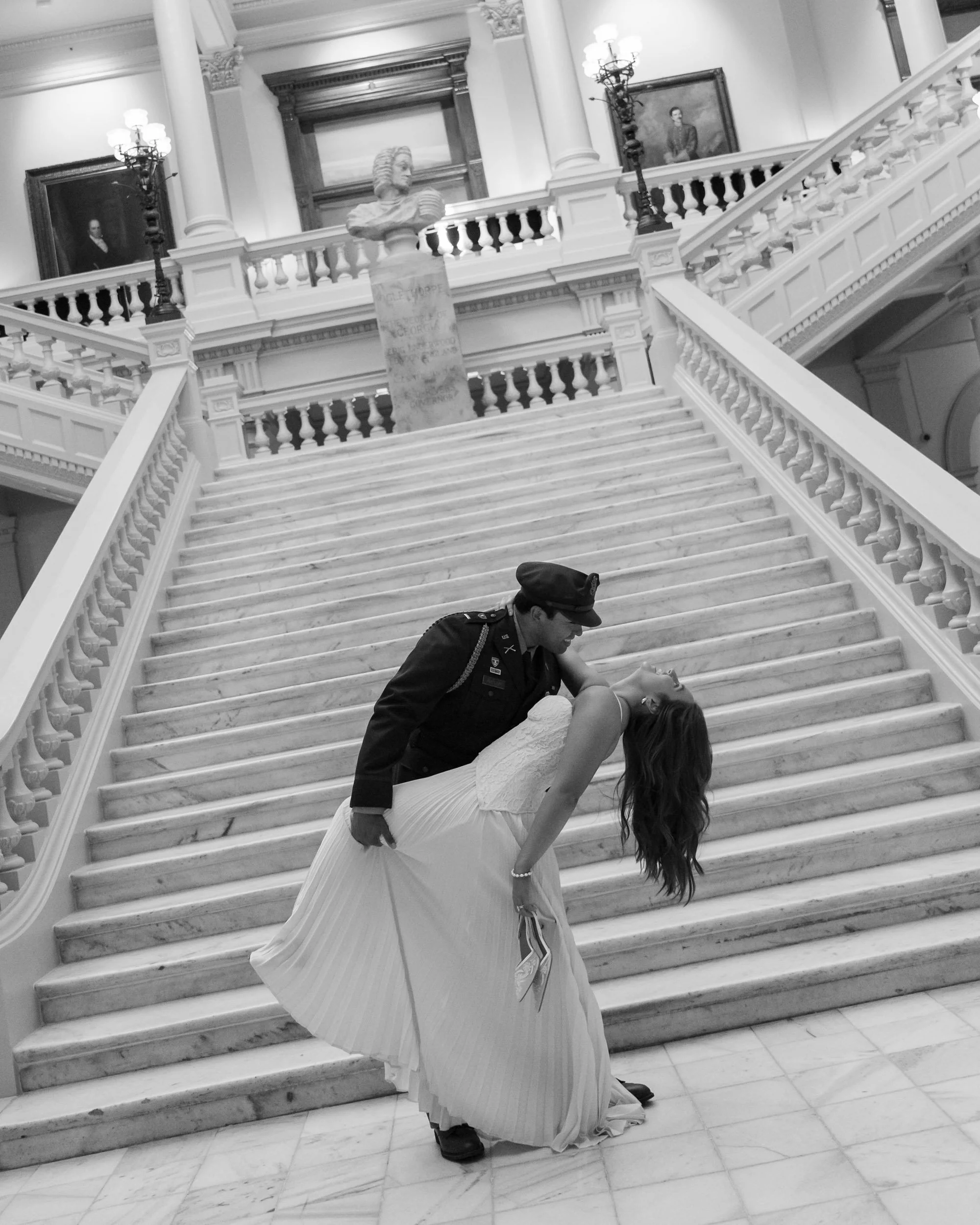 A soldier in uniform dips a woman in a wedding dress on a staircase inside a grand building with ornate railings and portrait paintings.