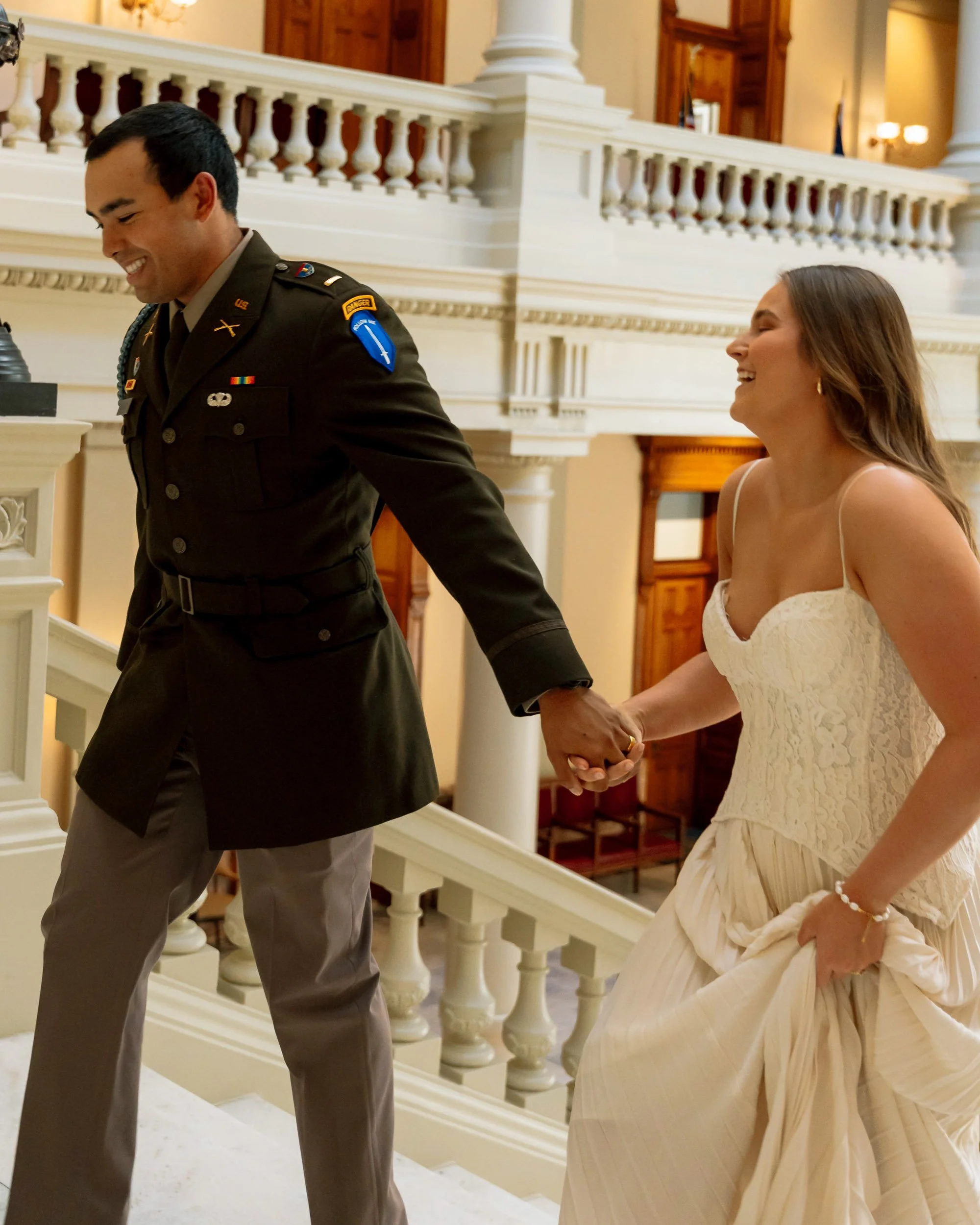 A happy couple holding hands on a staircase inside a building with ornate white railings and wooden paneling. The man is in military uniform, and the woman is in a white lace wedding dress.