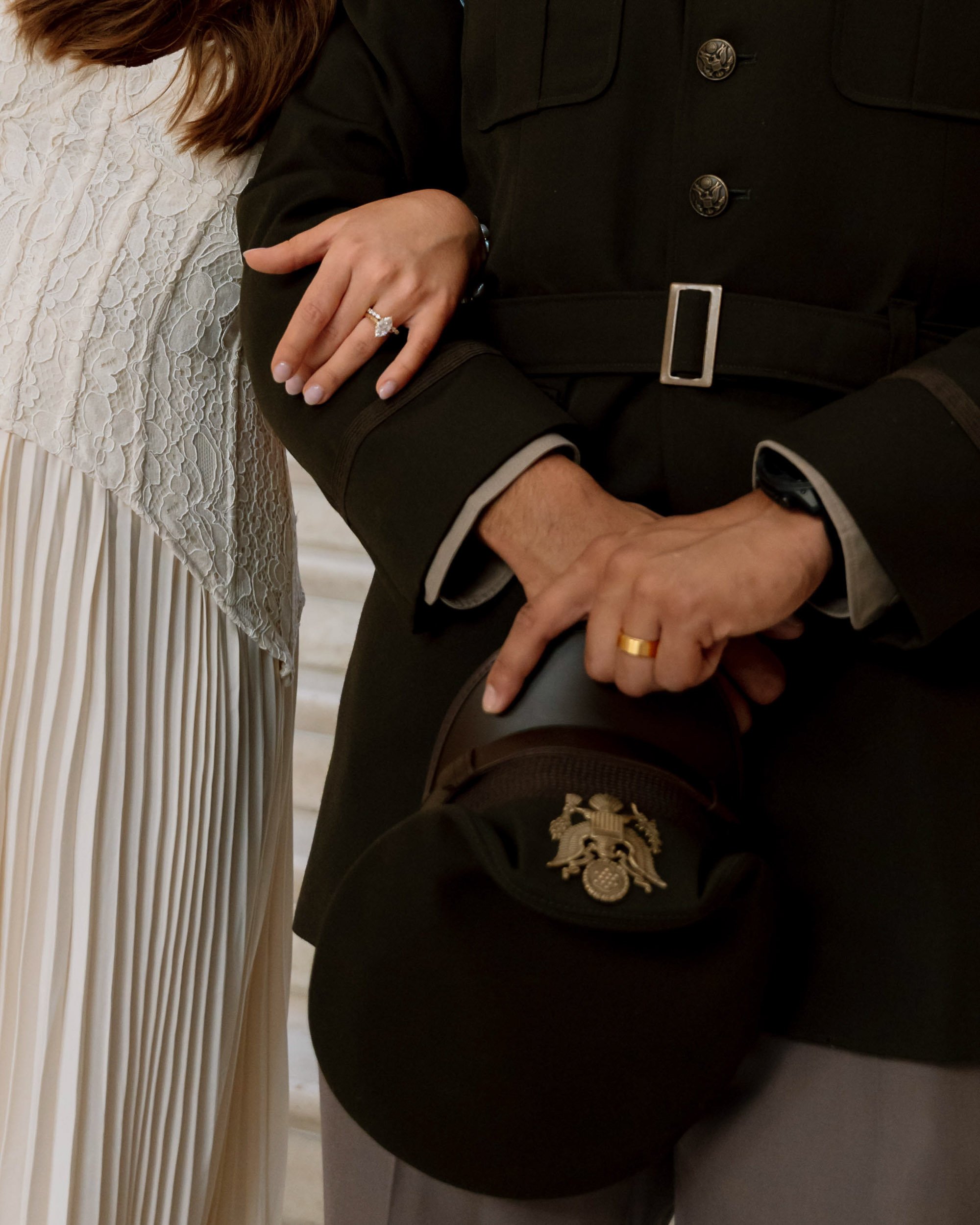 Close-up of a couple holding hands, with the woman's hand resting on the man's arm. They are both wearing wedding rings. The woman's hand has a diamond engagement ring and a wedding band. The man is wearing a military uniform with an insignia on his 