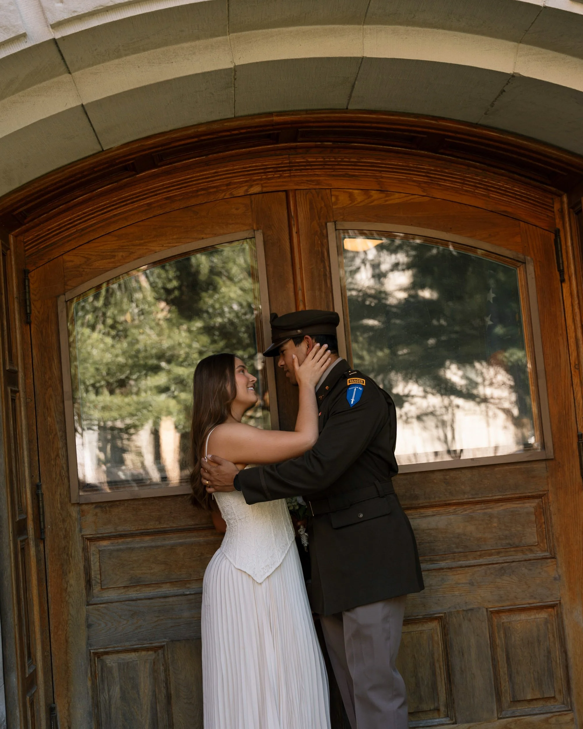 A bride and a groom in military uniform embrace and look into each other's eyes inside a wooden doorway with large windows, surrounded by greenery.