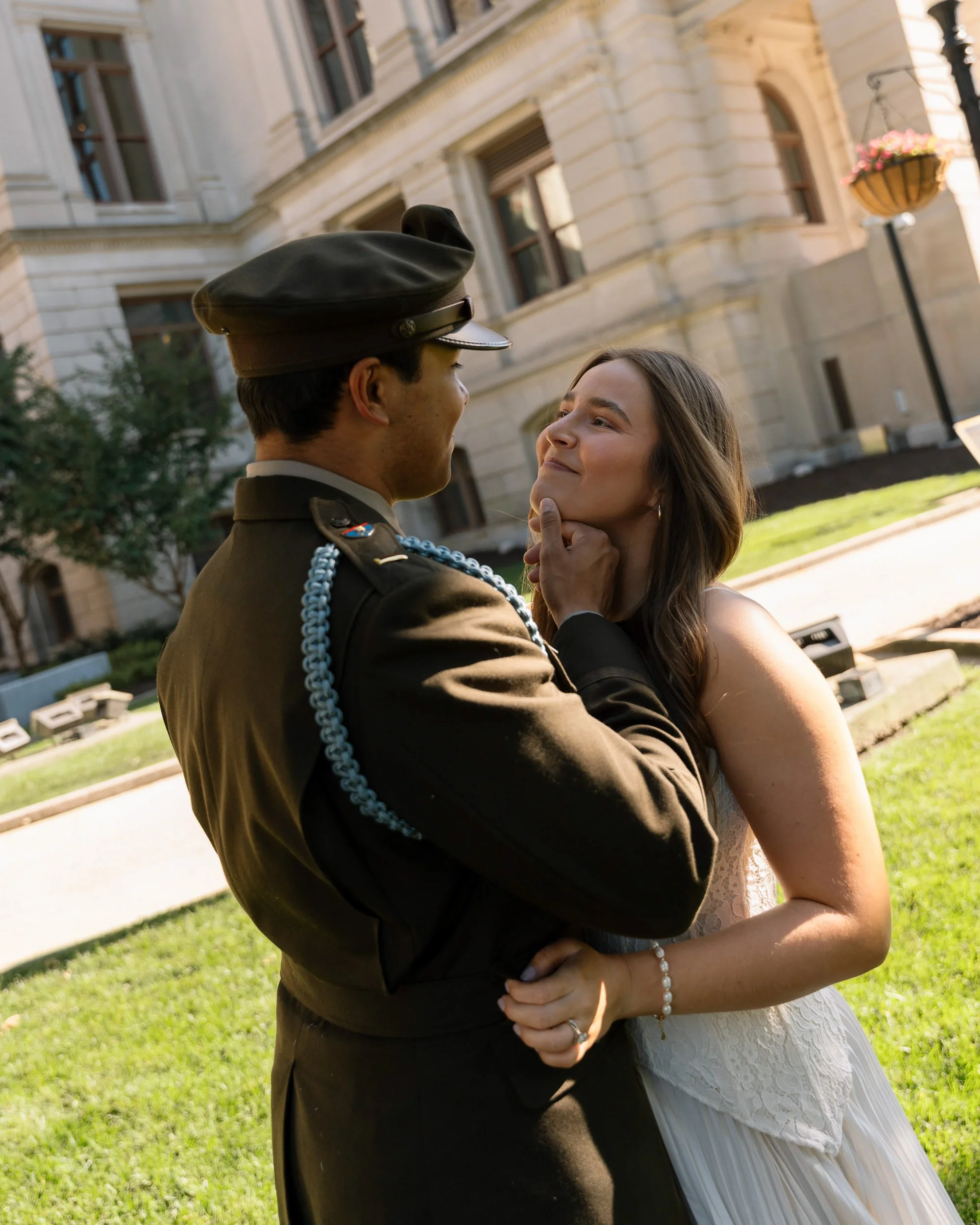A couple sharing a moment outdoors, with the woman gently touching the man's face, standing in front of a classical building with large windows, green grass, and hanging flower baskets.