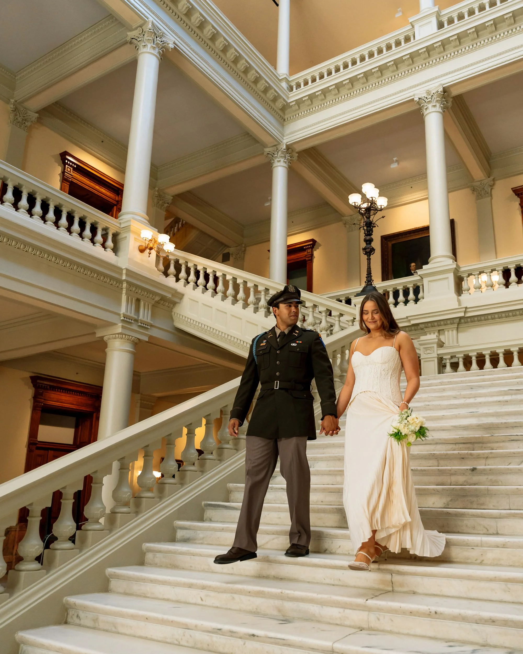 A bride in a wedding dress holding a bouquet and a groom in a military uniform holding hands, standing on a grand staircase inside a historic building with ornate columns and railings.