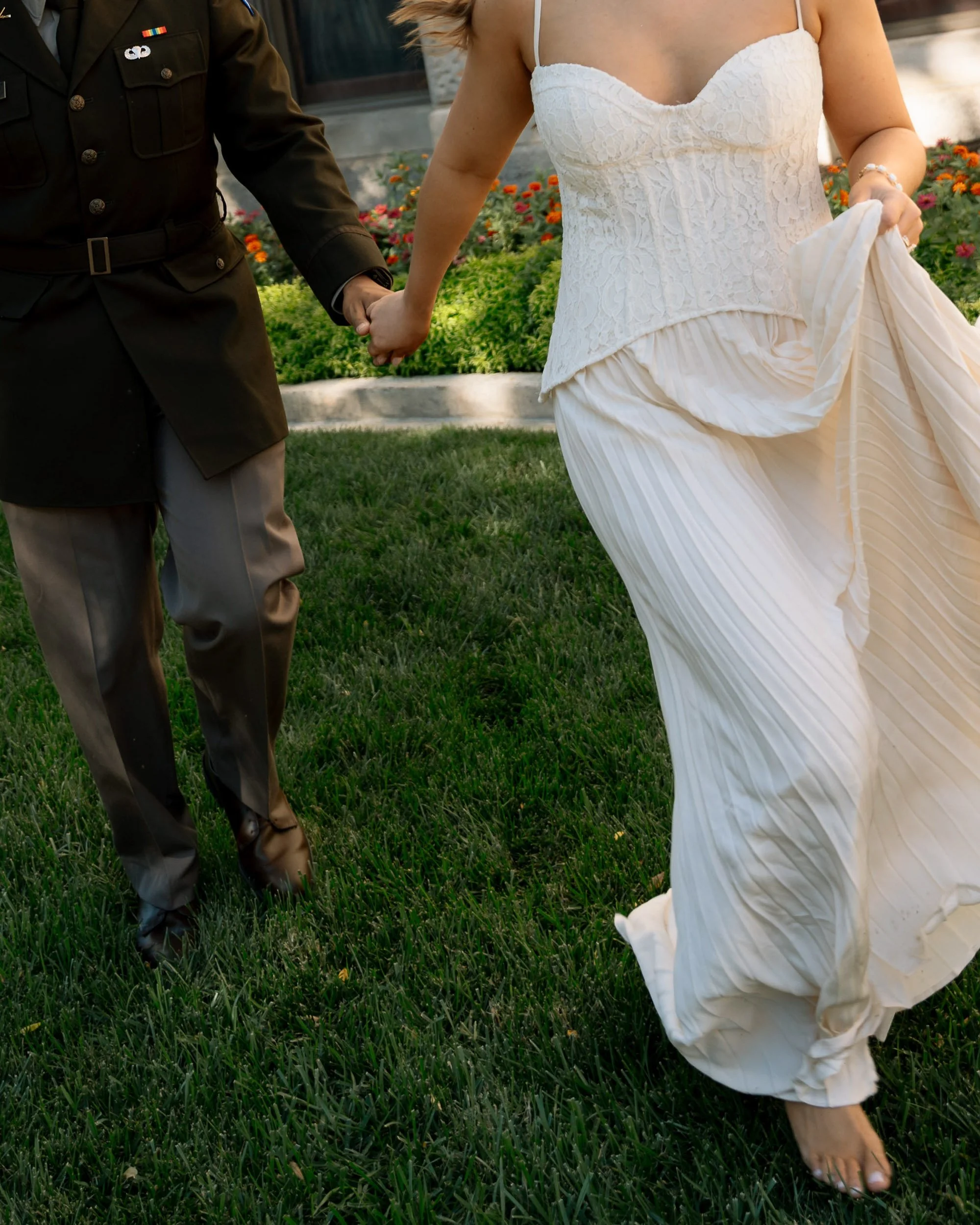 Couple holding hands in wedding attire walking on grass with flowers in background.