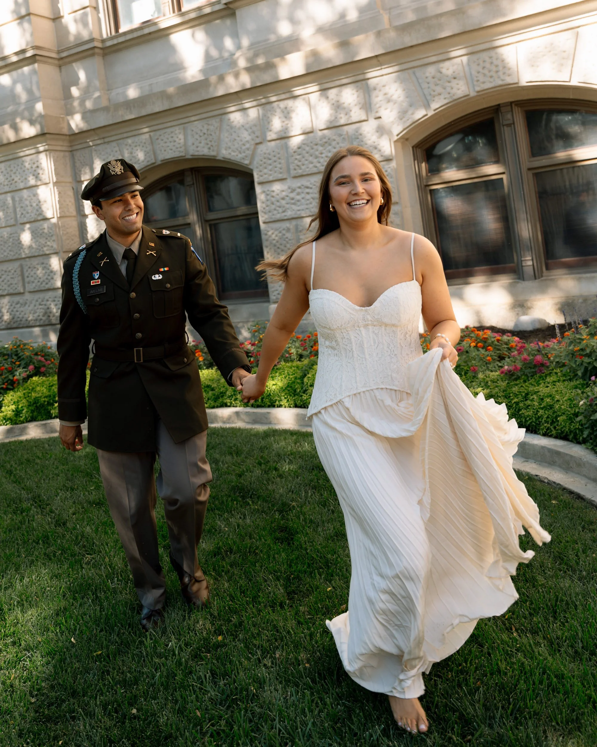 A man in a military uniform and a woman in a wedding dress holding hands and smiling, walking on grass in front of a stone building with flowers and large windows.