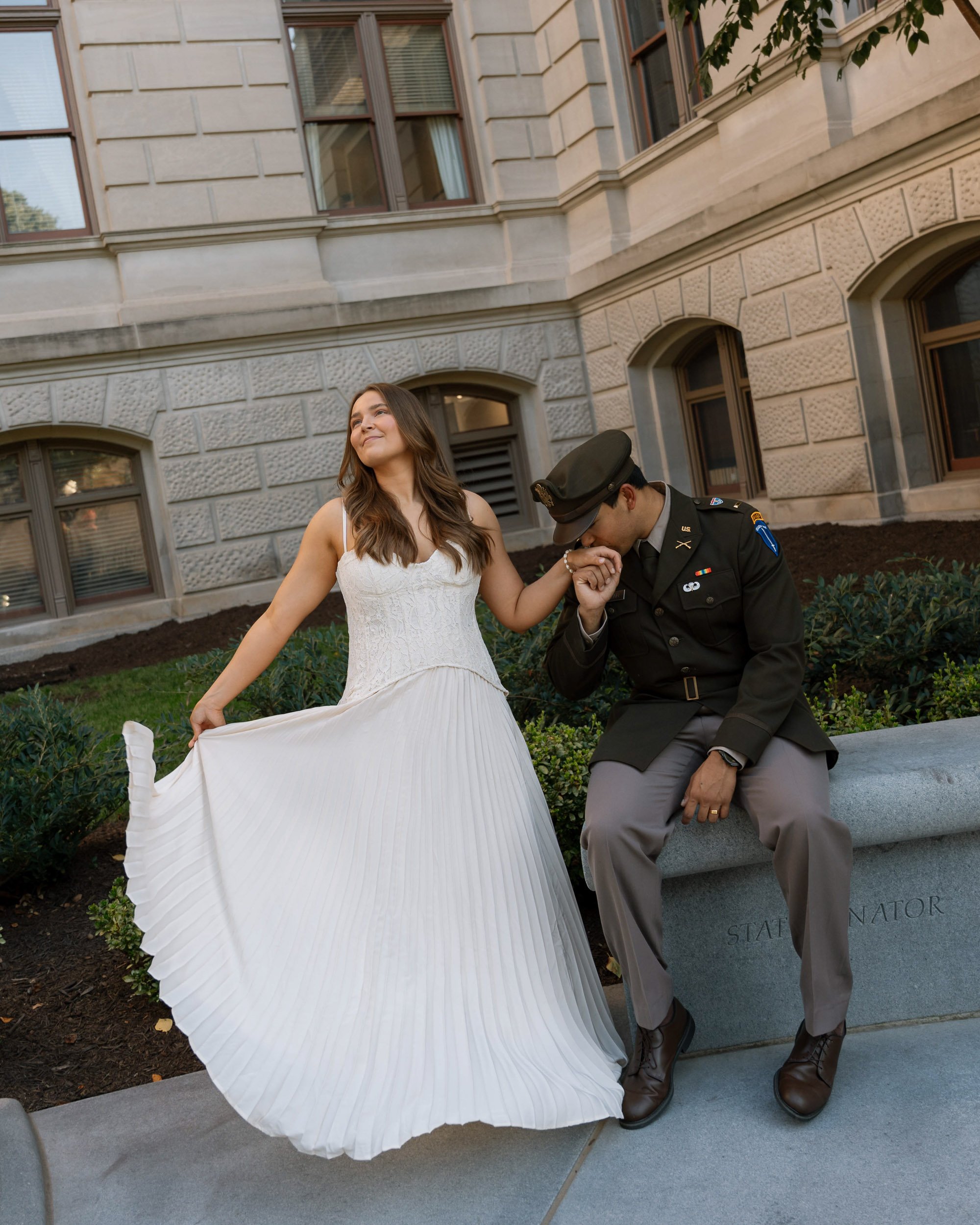 A woman in a white dress holds hands with a man in a military uniform while sitting on a stone bench outdoors near a government building.