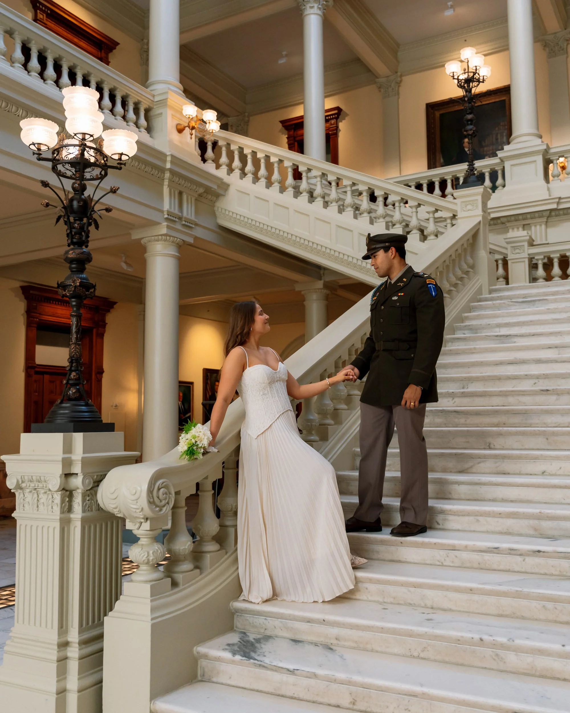 A bride in a white dress holding hands with a soldier in military uniform on a grand marble staircase inside a building with ornate decor.