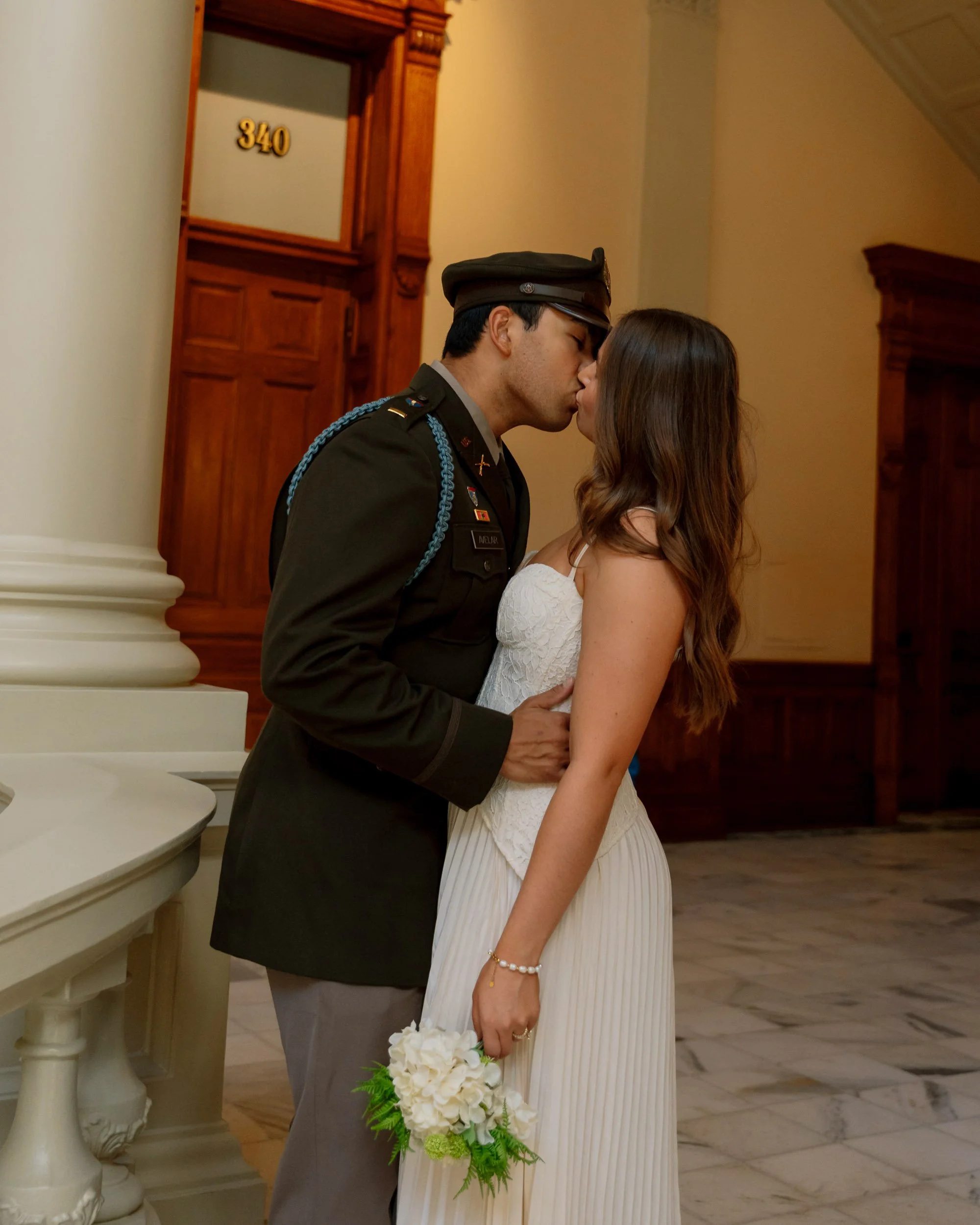 A couple kissing, the man is in a military uniform and the woman is holding a bouquet of white flowers, inside a building with wooden decor and marble flooring.