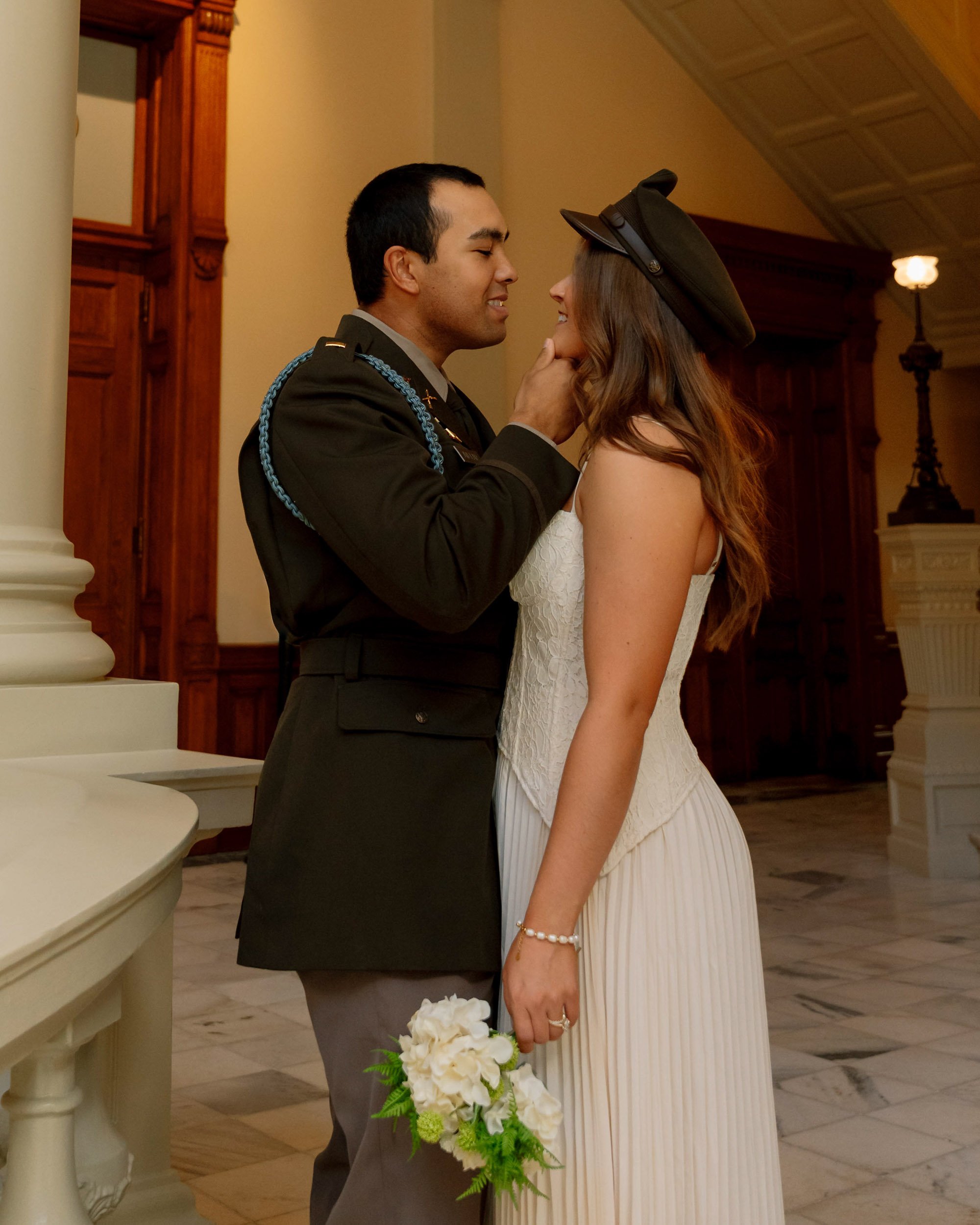 A couple, a man in a military uniform and a woman in a white dress, about to kiss in an indoor setting, with a woman holding a bouquet of white flowers.