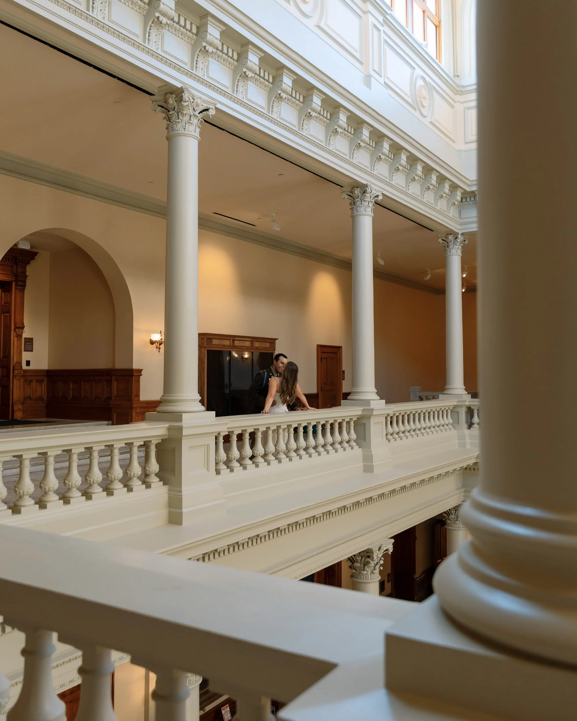A man and woman standing close together on an elegant balcony with white railings, in a grand building with tall columns and intricate decorative molding, during daytime.