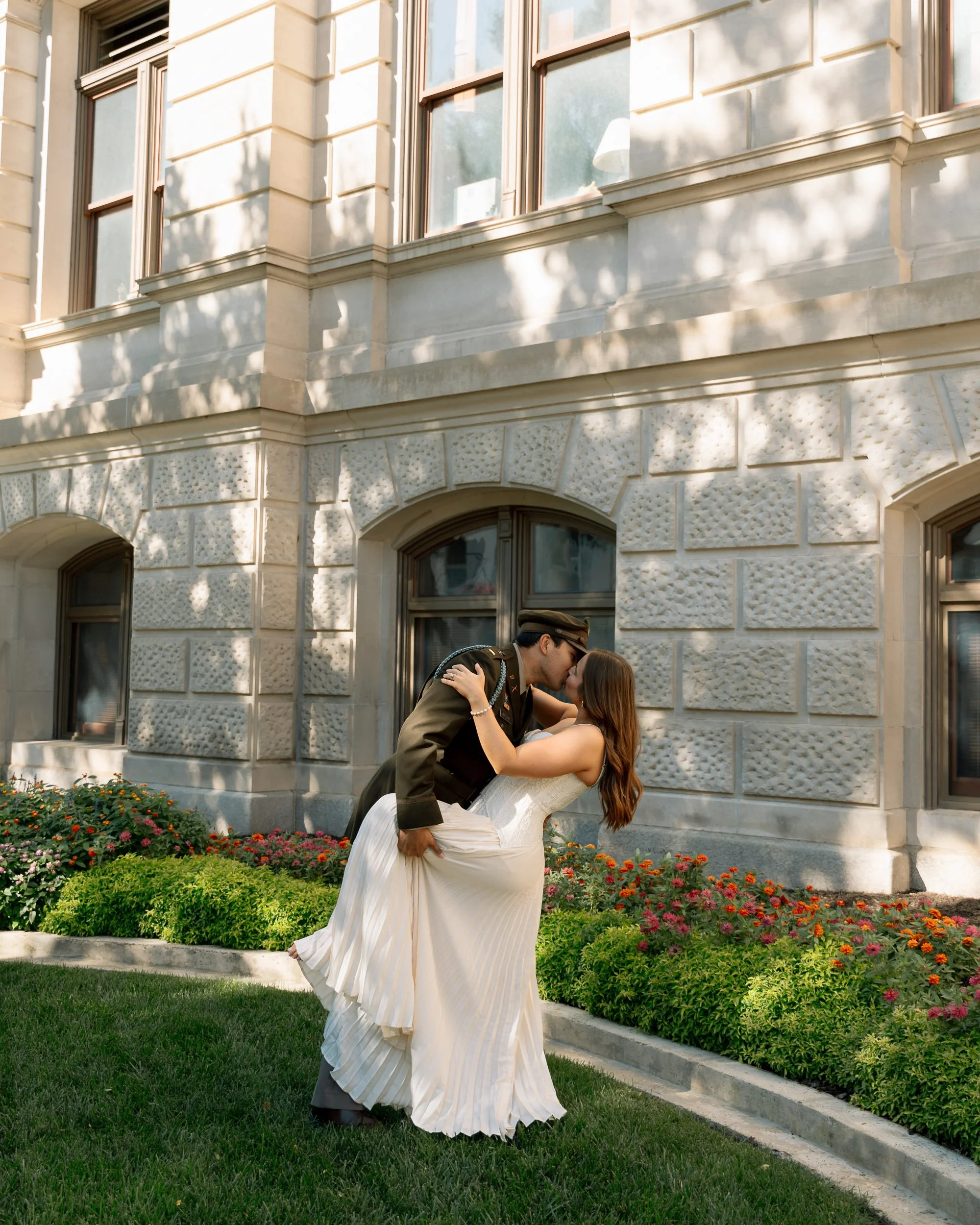 A couple in wedding attire sharing a kiss outdoors in front of a stone building with flower beds.