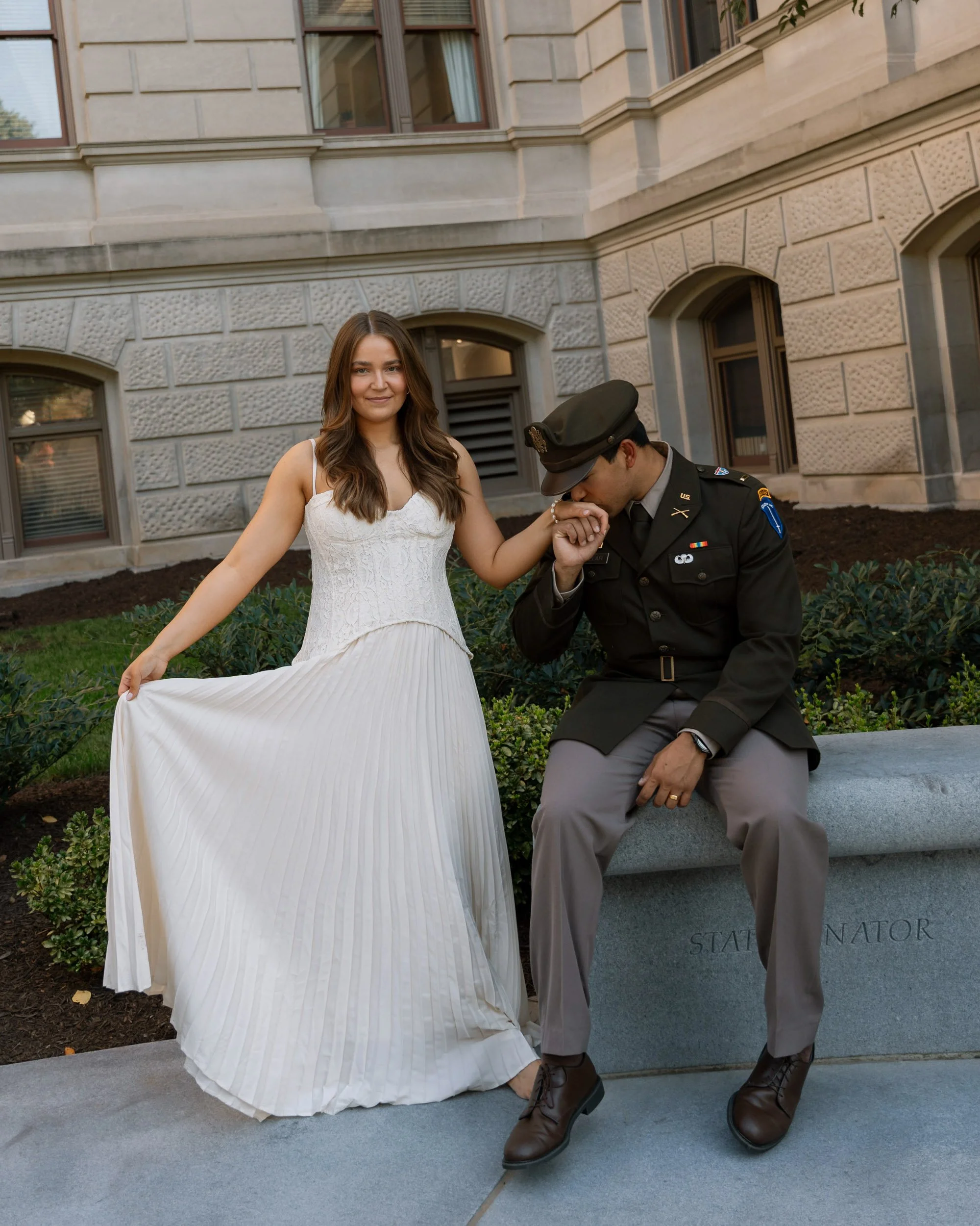 A woman in a white dress holding a man's hand as he kisses it. The man is dressed in a military uniform and is sitting on a stone bench outside a stone building with large windows.