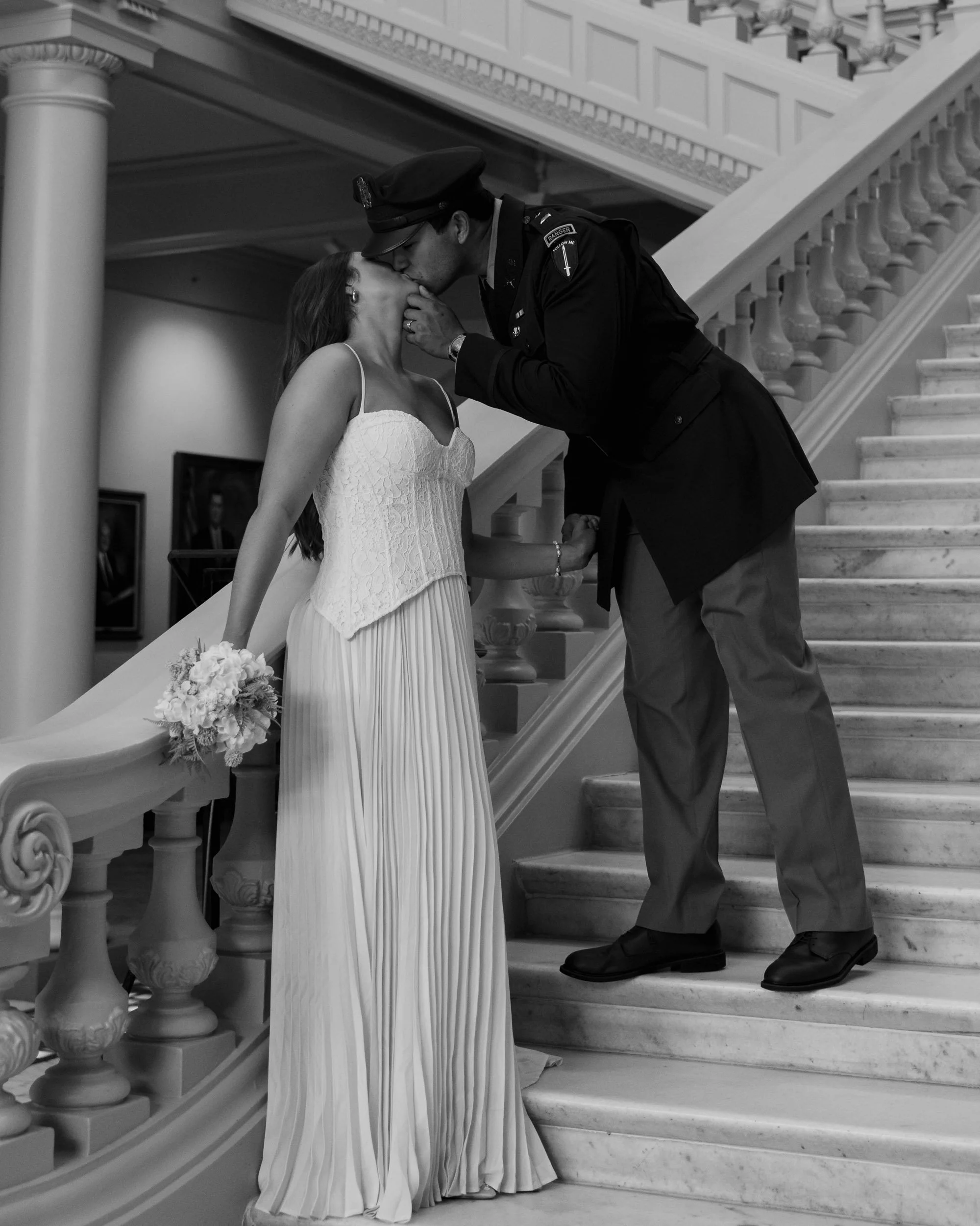 A couple in wedding attire sharing a kiss on a staircase, with a man in a military uniform and a woman in a wedding dress holding a bouquet.