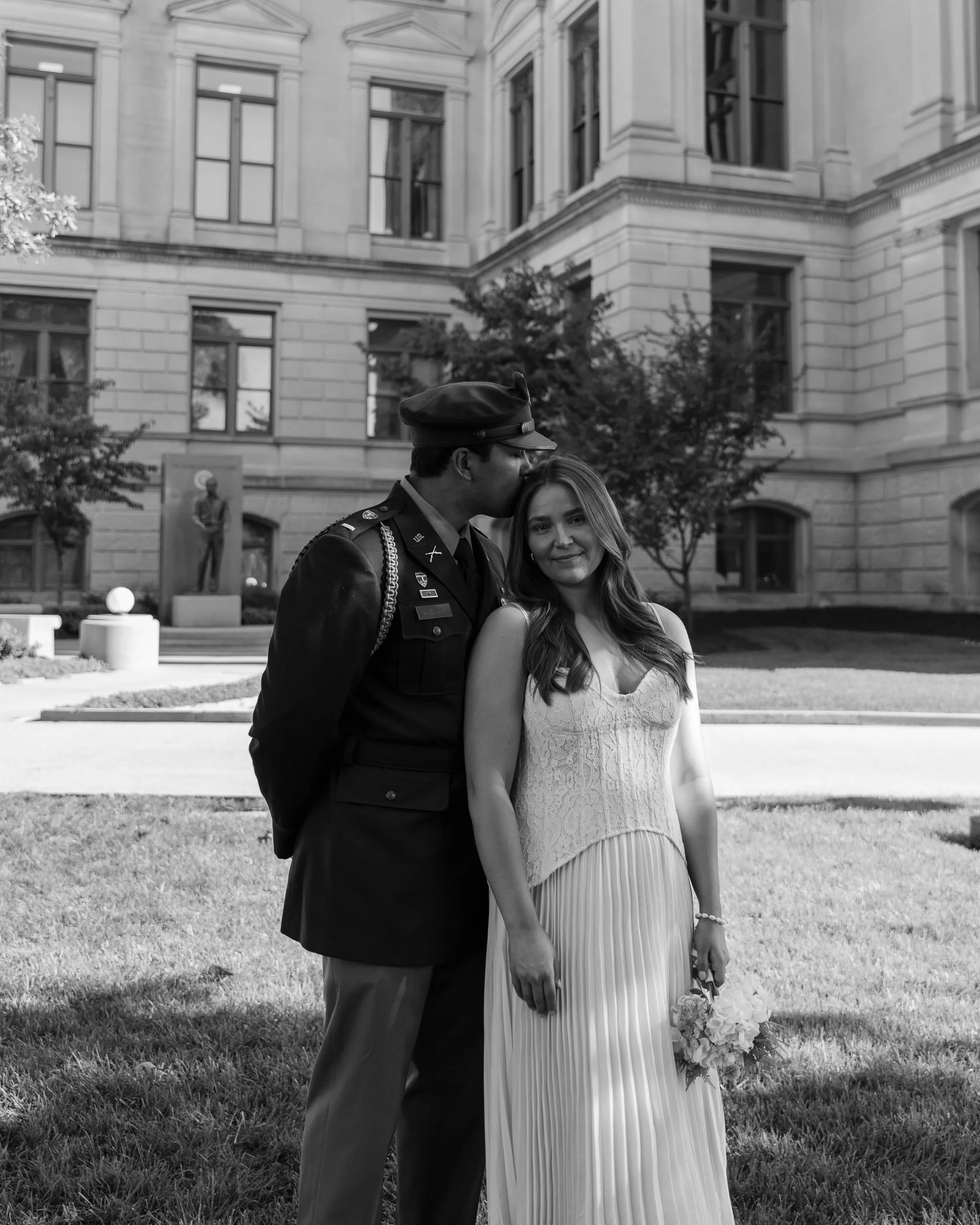 A soldier kisses a young woman on the forehead in front of a historic building, with the woman holding a bouquet of flowers, in black and white.