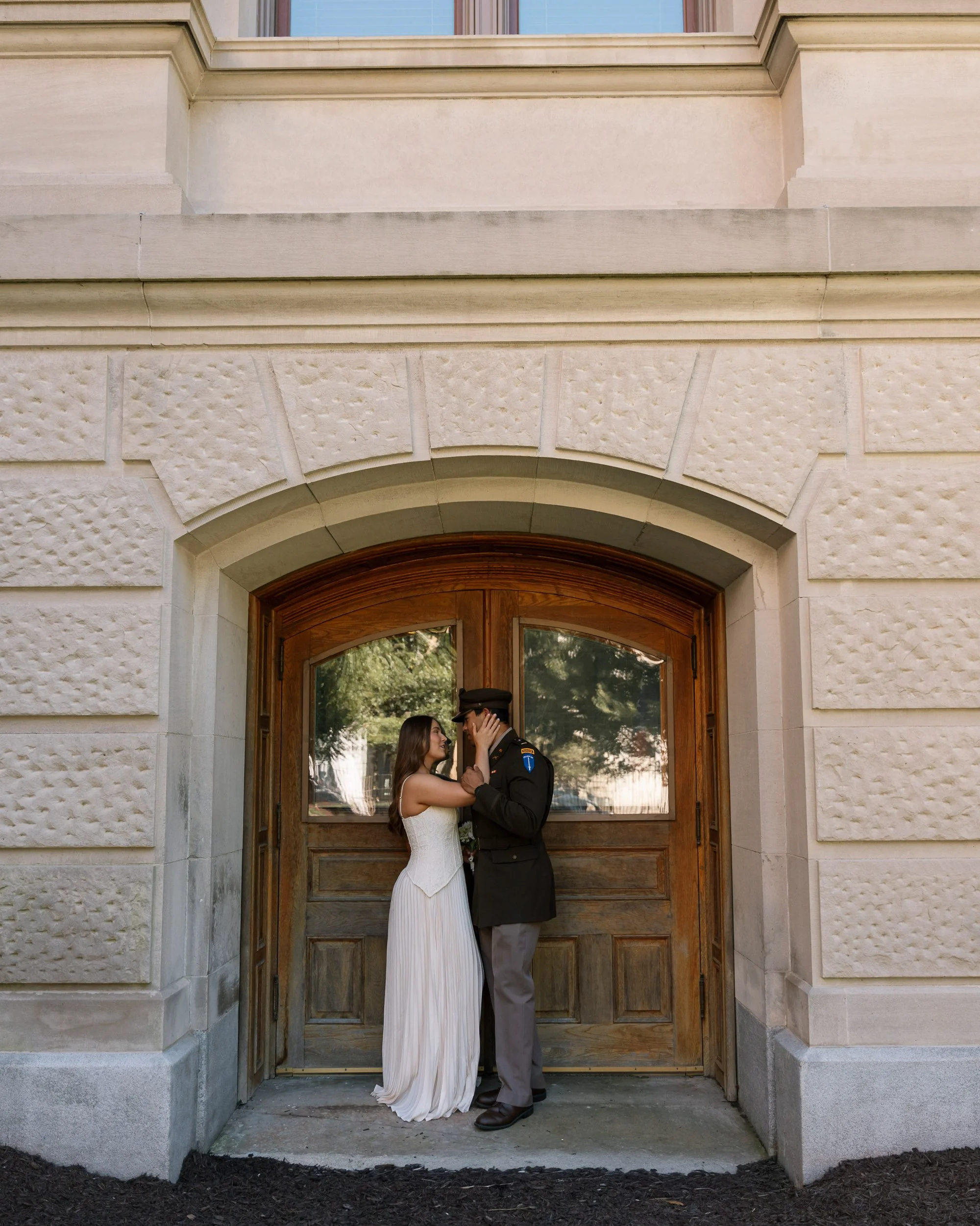 A couple in wedding attire sharing a kiss in front of a wooden door on a building with stone exterior walls.