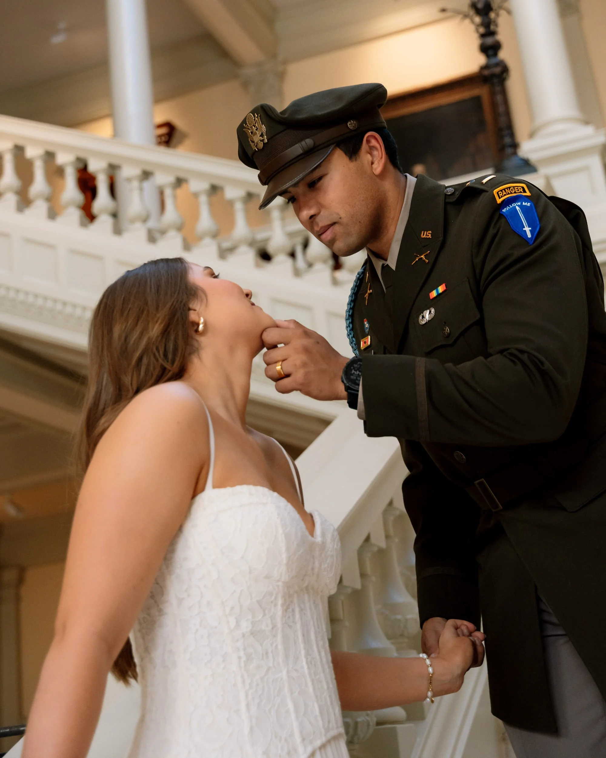 A woman in a white dress having her chin gently touched or kissed by a man in a military uniform inside a building with ornate architecture.