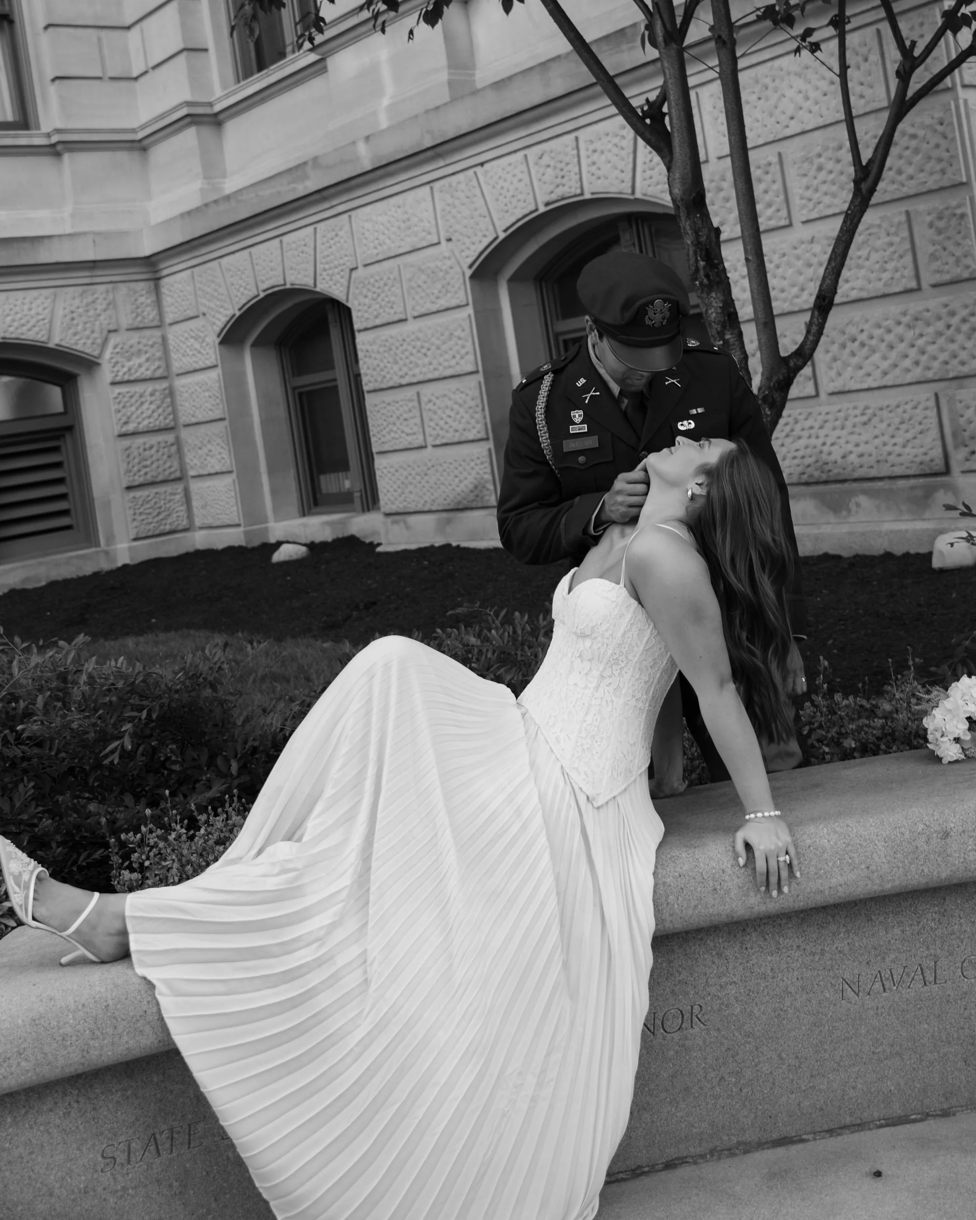 A woman in a wedding dress sitting on a stone ledge, leaning back with her head tilted up, as a man in a military uniform leans over her, appearing to kiss her or whisper to her. They are outdoors in front of a building with large windows and a tree.