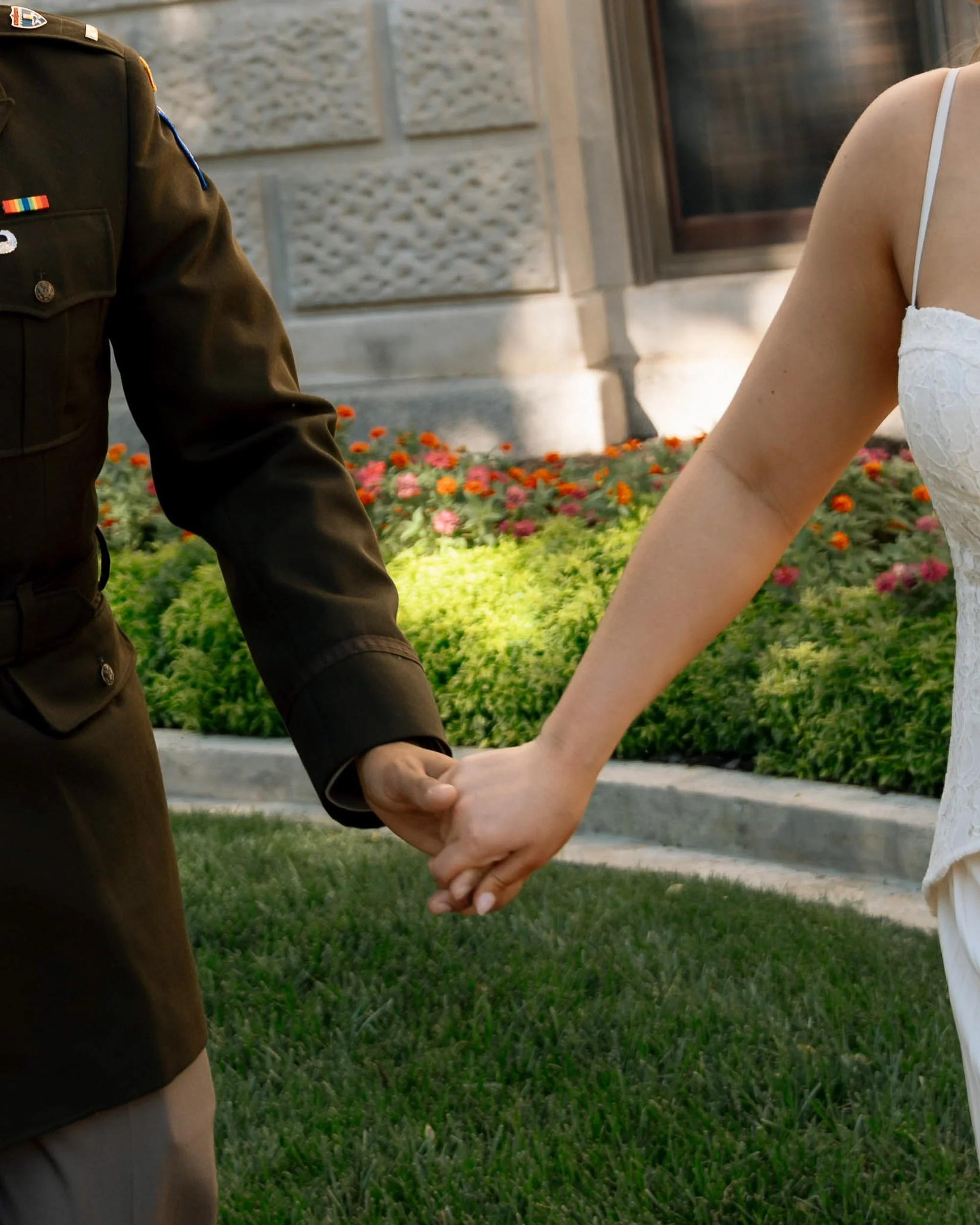 A person in a military uniform holding hands with a person in a white dress outdoors in front of a building and flower bed.