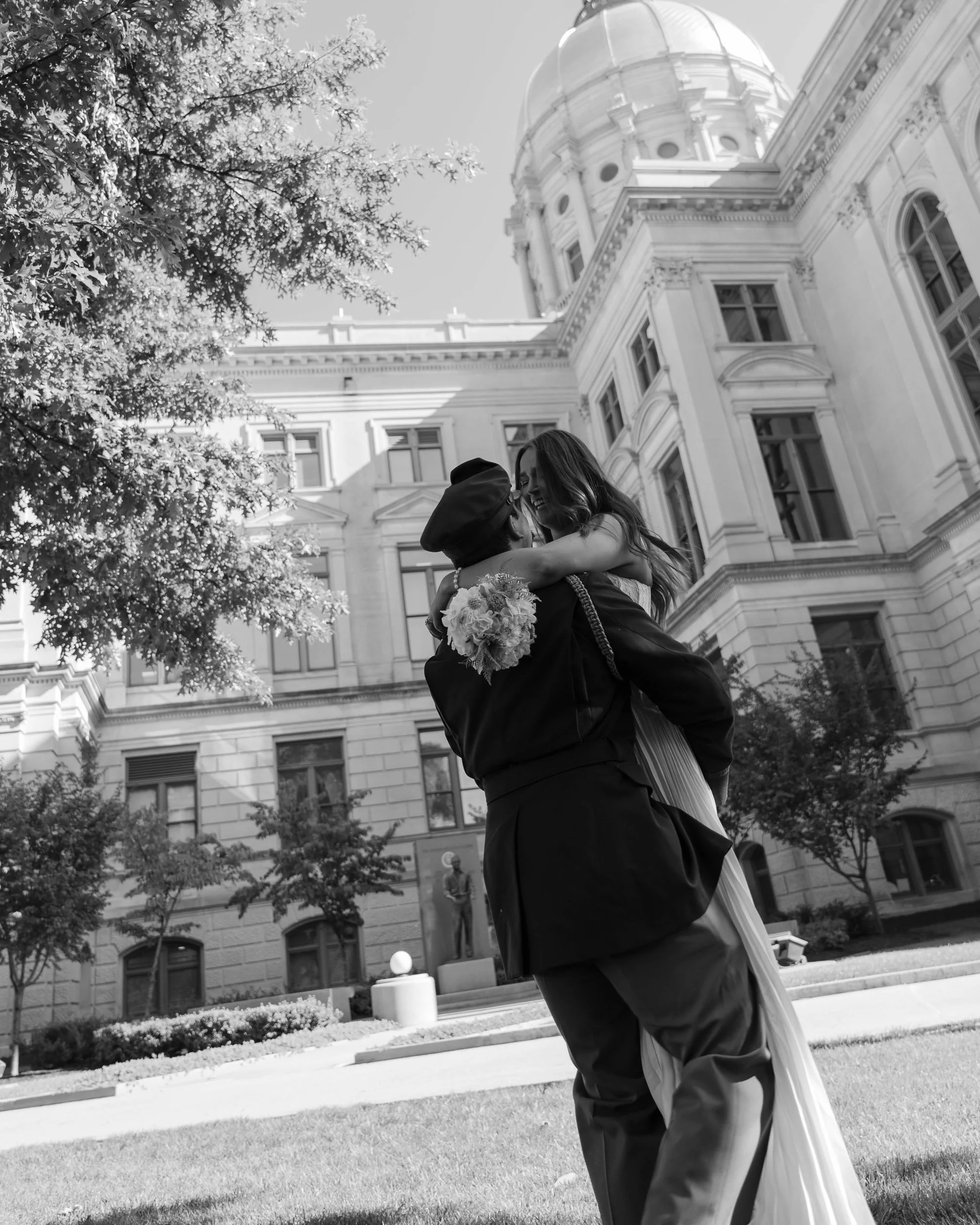 A couple, one holding a bouquet, embraces outside a historic building with architectural details, trees, and a sculpture in the background.