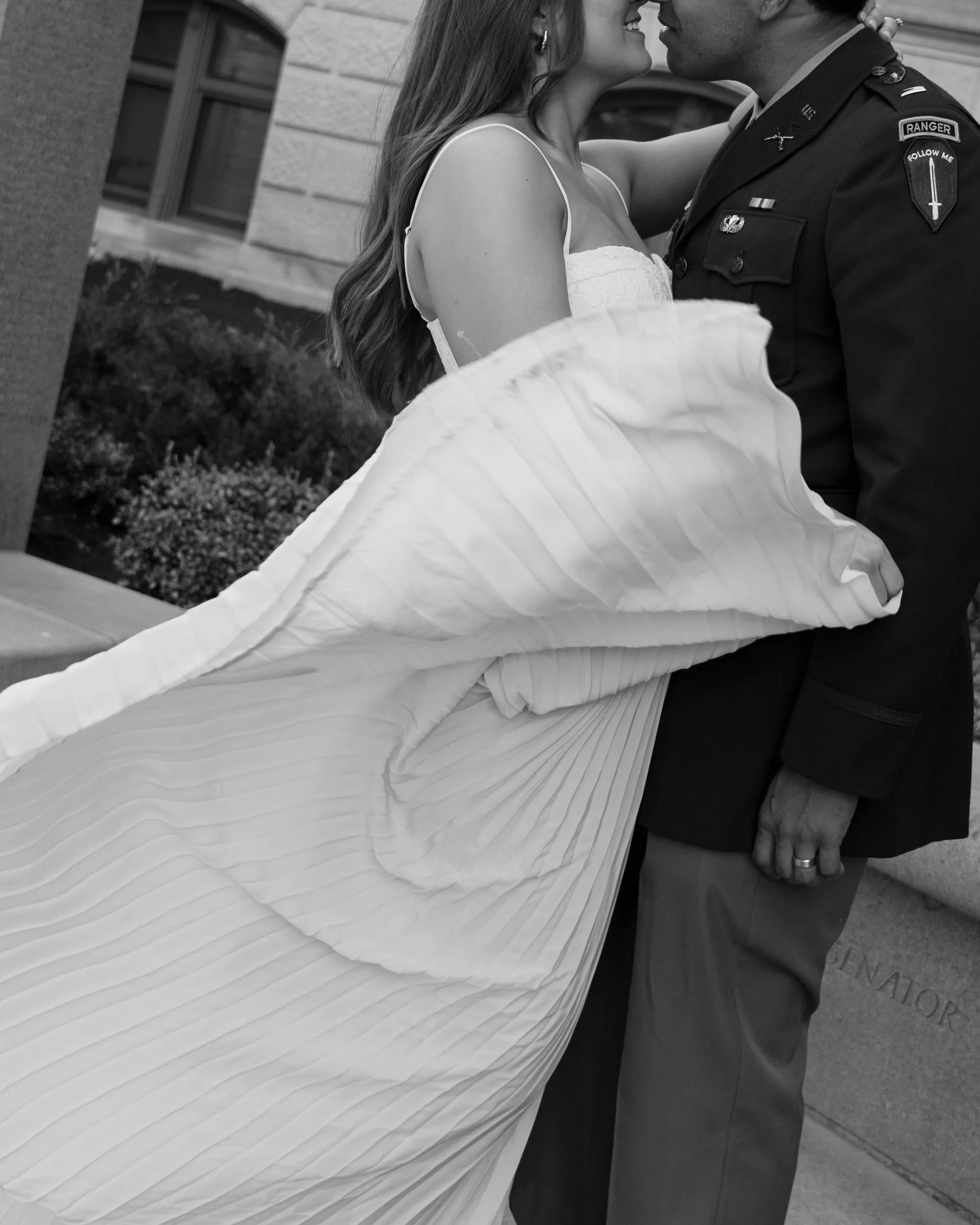 A bride and a man in military uniform are about to kiss, with the bride holding her wedding dress.