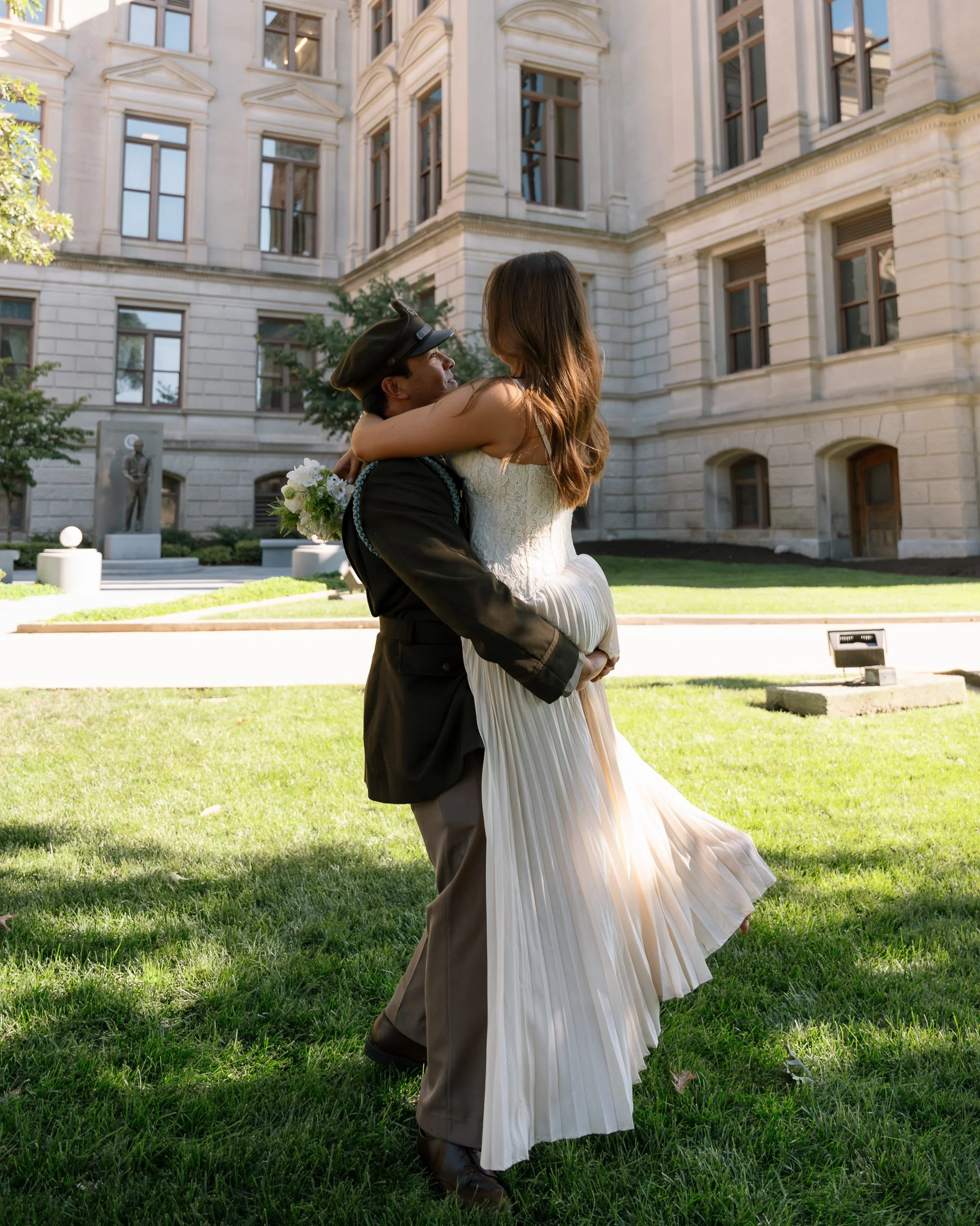 A man in uniform holding a woman in a white dress in a garden in front of a historic building.