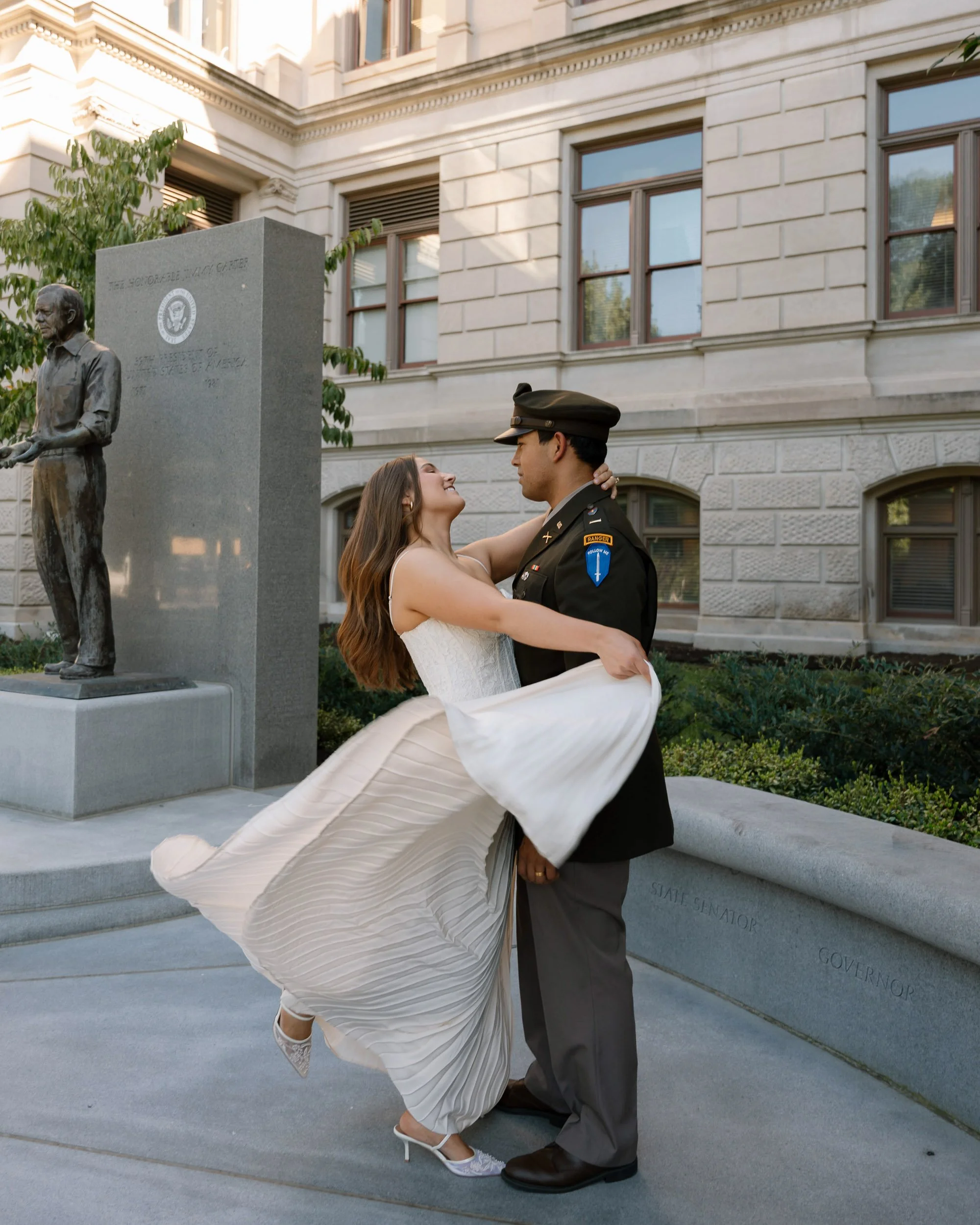 A woman in a white dress and heels being lifted by a man in a military uniform, smiling at each other outdoors in front of a sculpture and a stone building.
