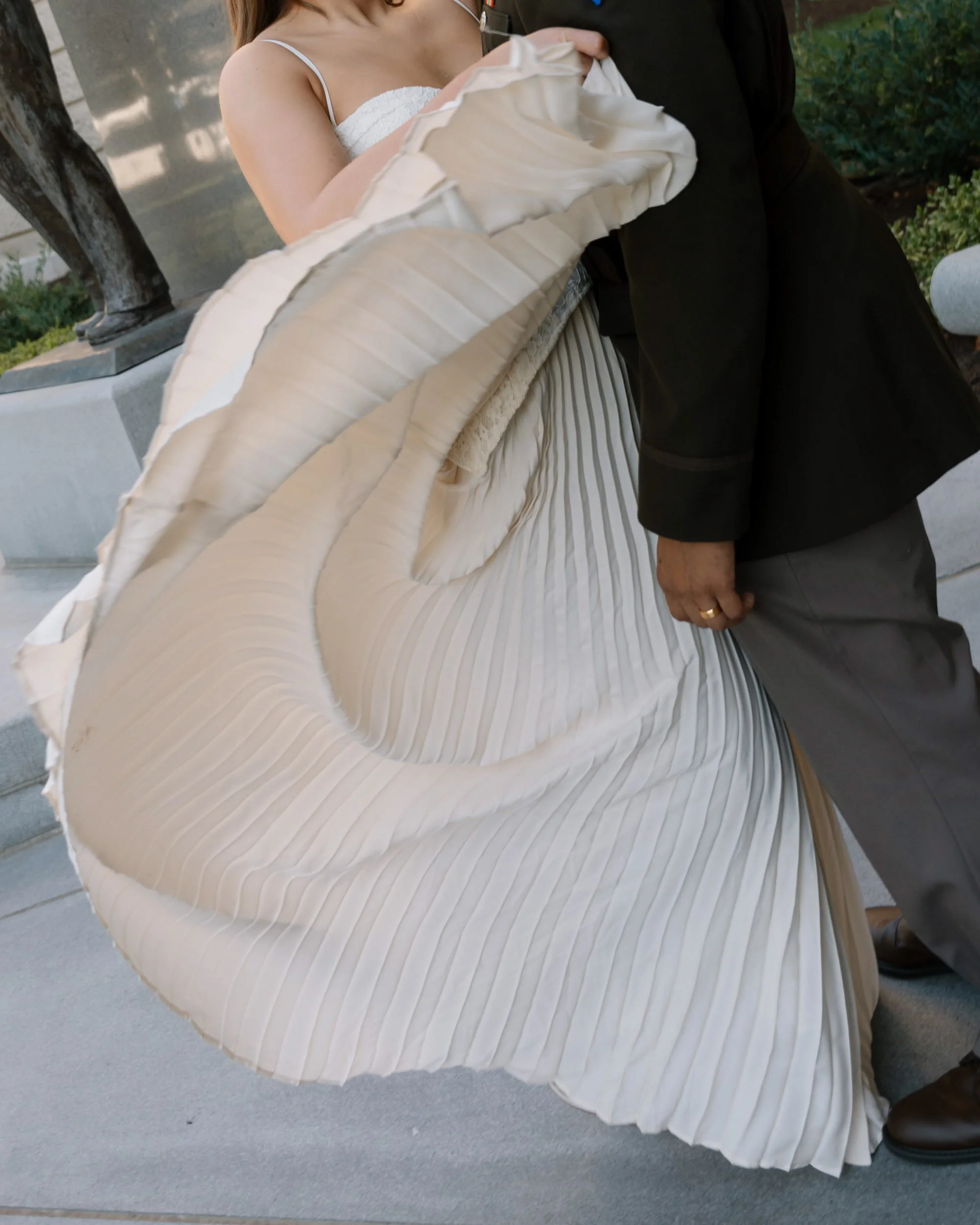 Close-up of a woman wearing a white dress with pleated details, possibly at a wedding or formal event, with a man dressed in dark clothing.