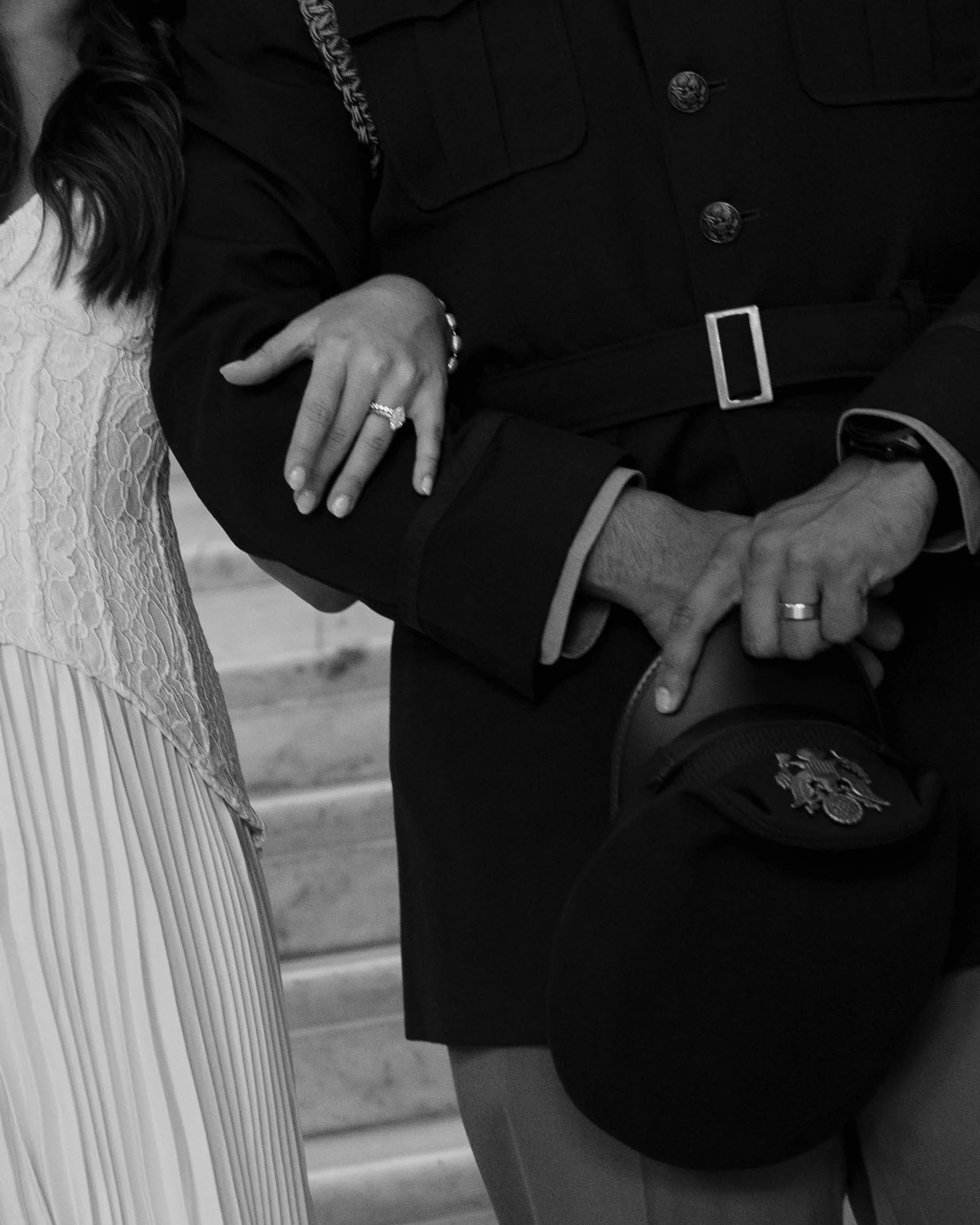 A black and white photo of a man in a police uniform holding hands with a woman wearing a ring and a lace top. The woman’s hand rests on the man’s arm, and the man is pointing to his police badge with his other hand.