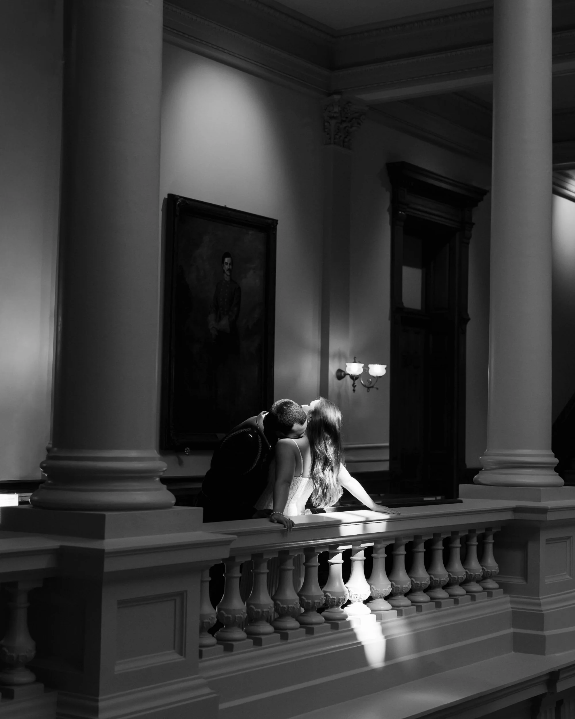 A black and white photo of a couple kissing on an elegant balcony inside a grand building, with shadows from a window casting light onto the railing. Portrait paintings and decorative wall sconces decorate the background.