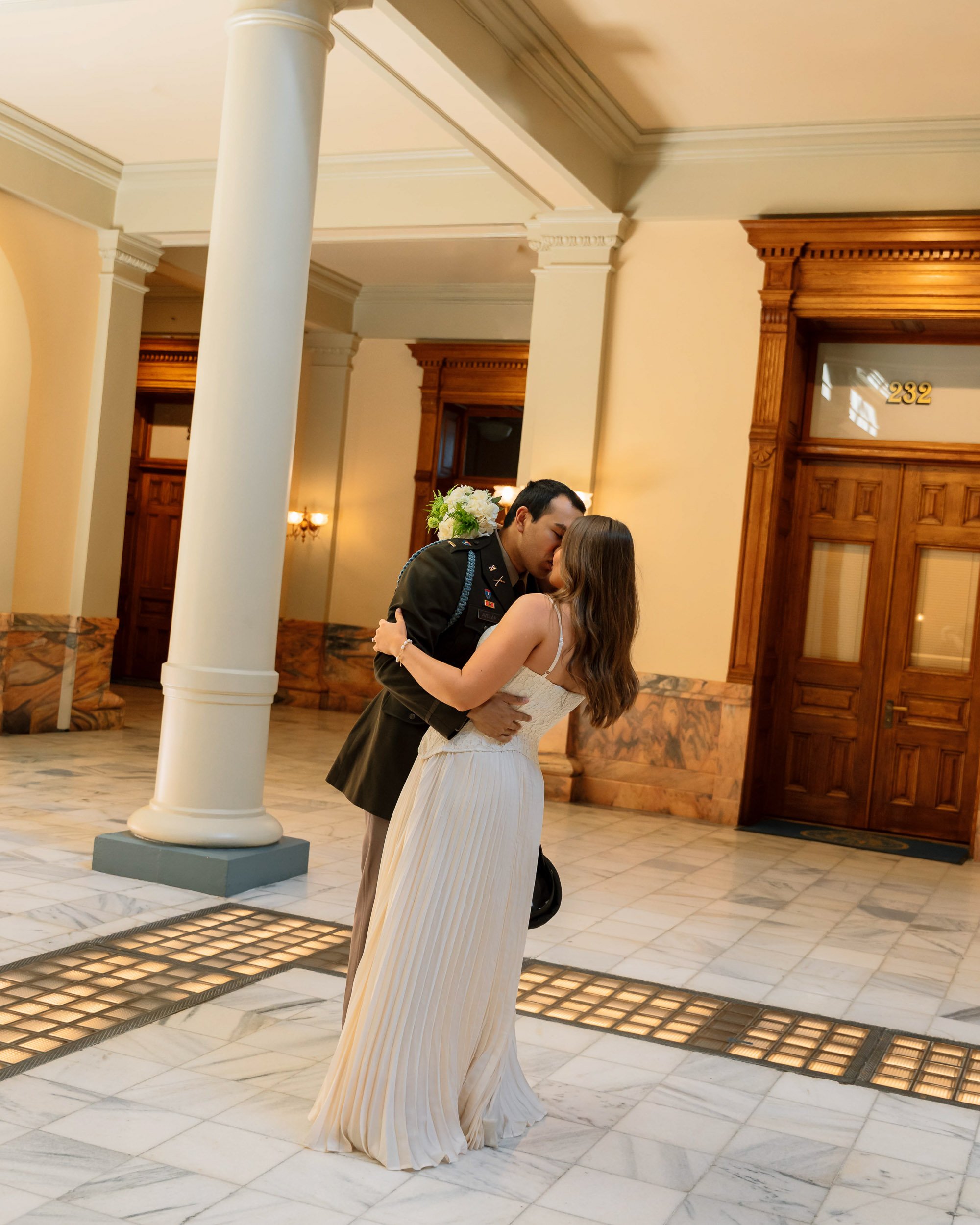 A couple kissing in an elegant indoor setting with marble floors, wooden doors, and columns. The woman is wearing a white pleated dress and the man is in a military uniform. The woman is holding a bouquet of flowers.
