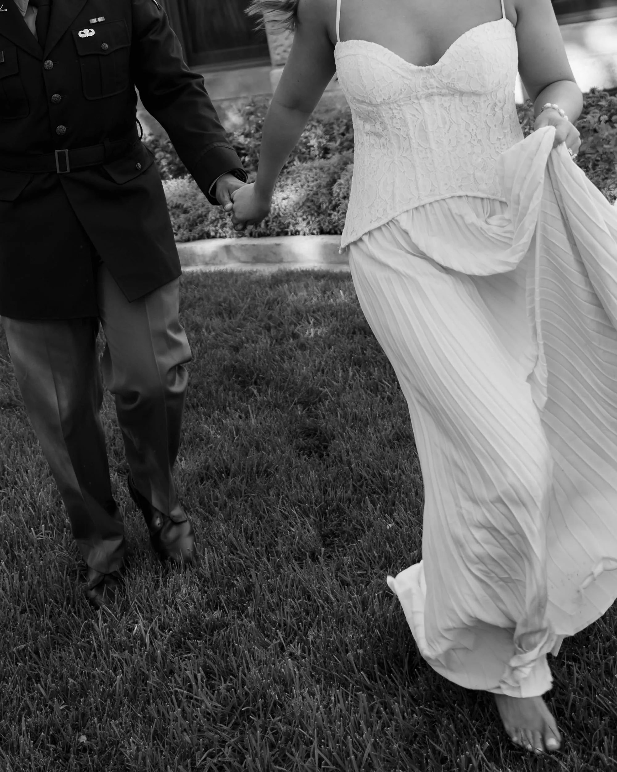 A bride and groom holding hands, walking on grass outdoors, bride wearing a strapless lace dress, groom in a military uniform.