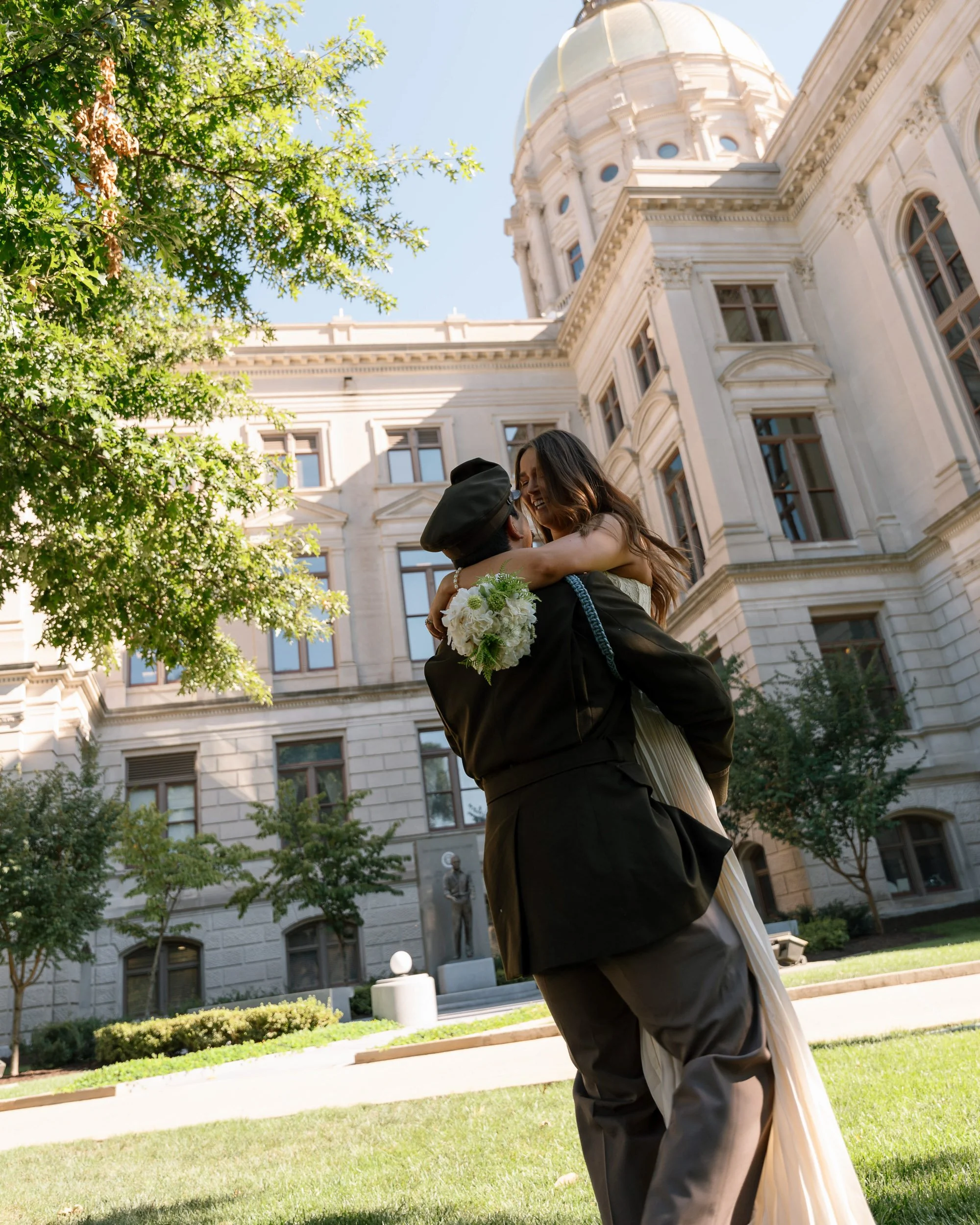 A couple, dressed in black and white, embraces and joyfully dances outside a historic white stone building with a large dome. The woman holds a bouquet of white flowers. Green trees and a manicured lawn surround them on a sunny day.