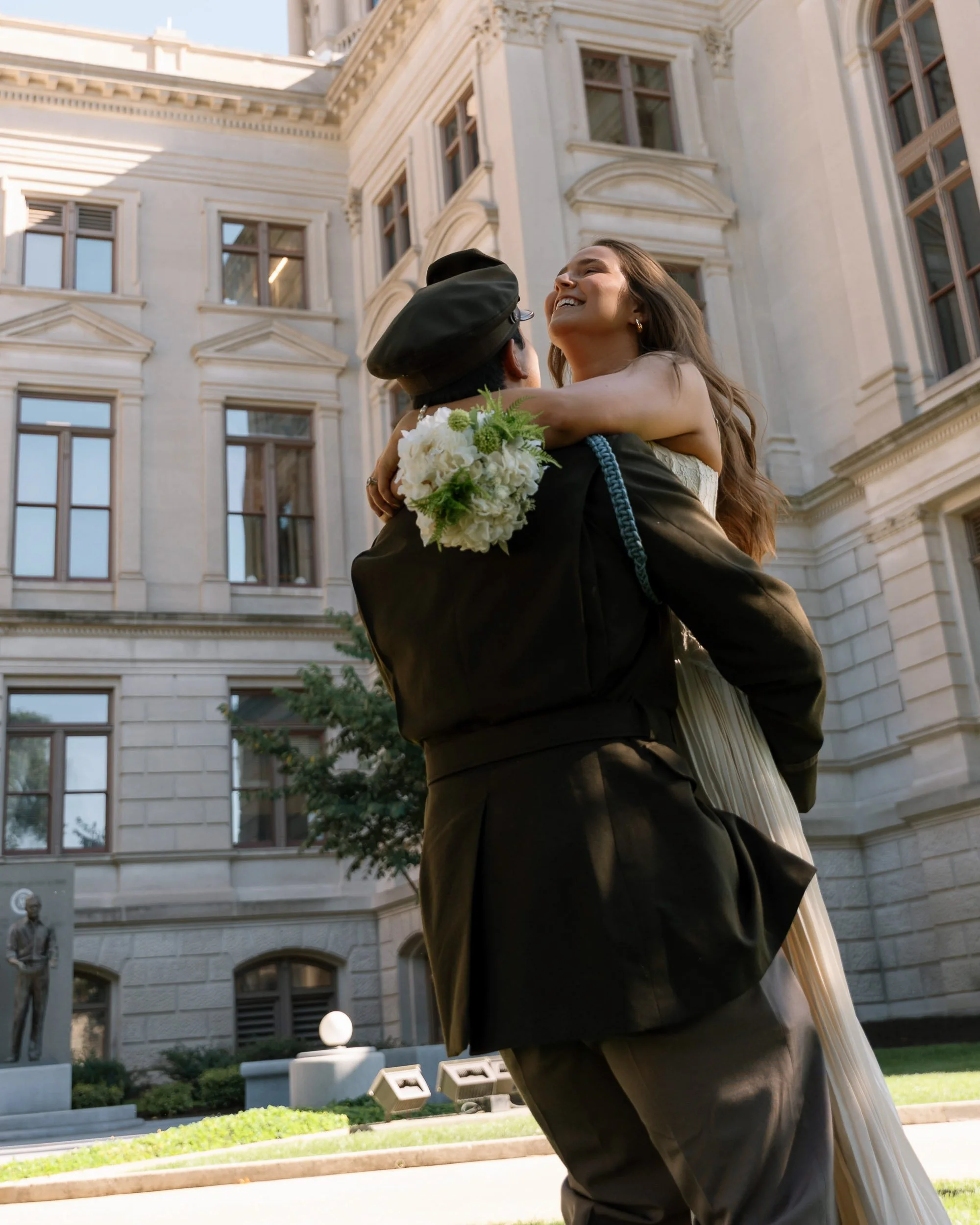 A woman in a white dress being lifted by a man in a military uniform, holding a bouquet of white flowers, outside a large historic building with tall windows.