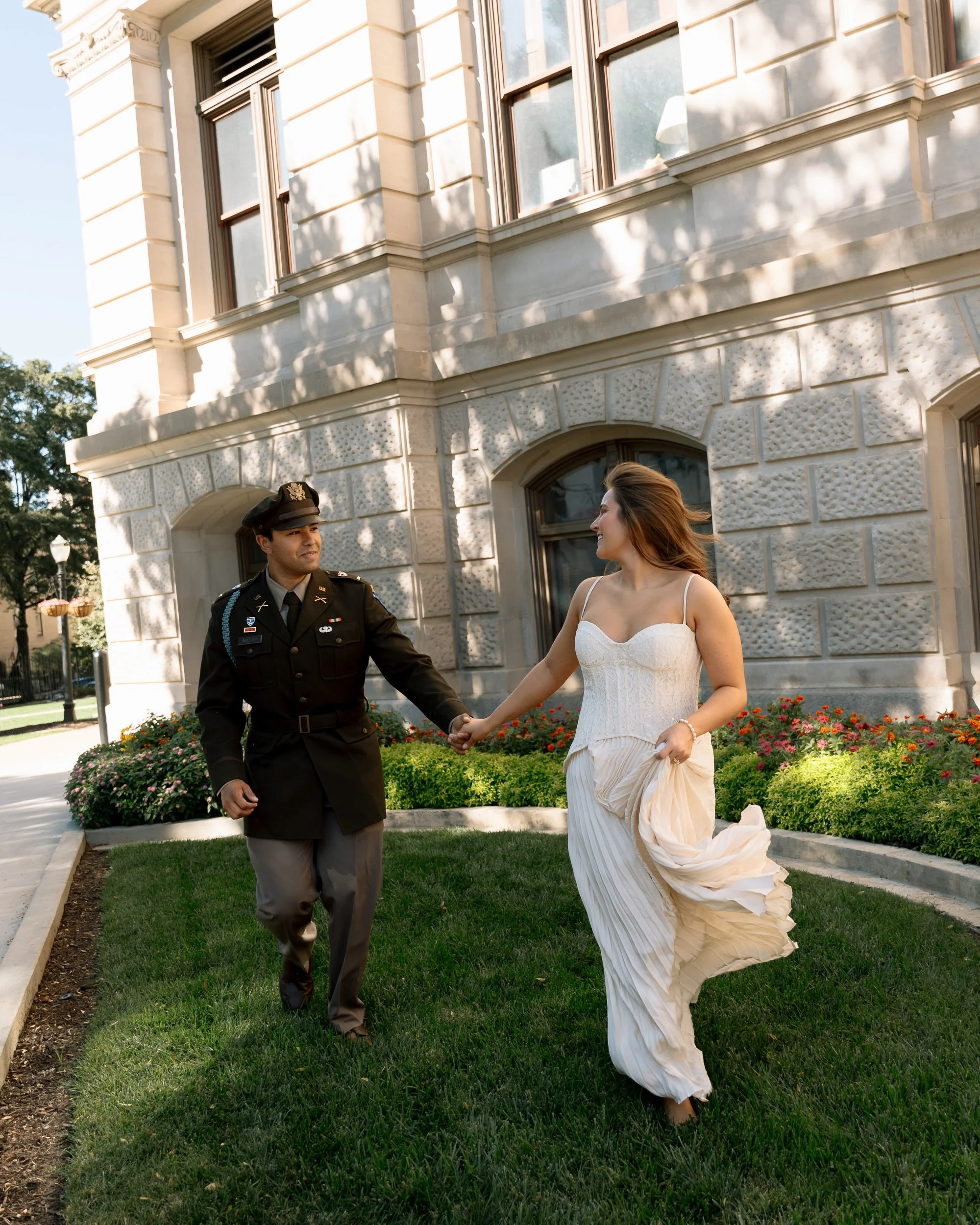 A woman in a white dress holding hands with a man in a uniform outside a stone building with flower beds.