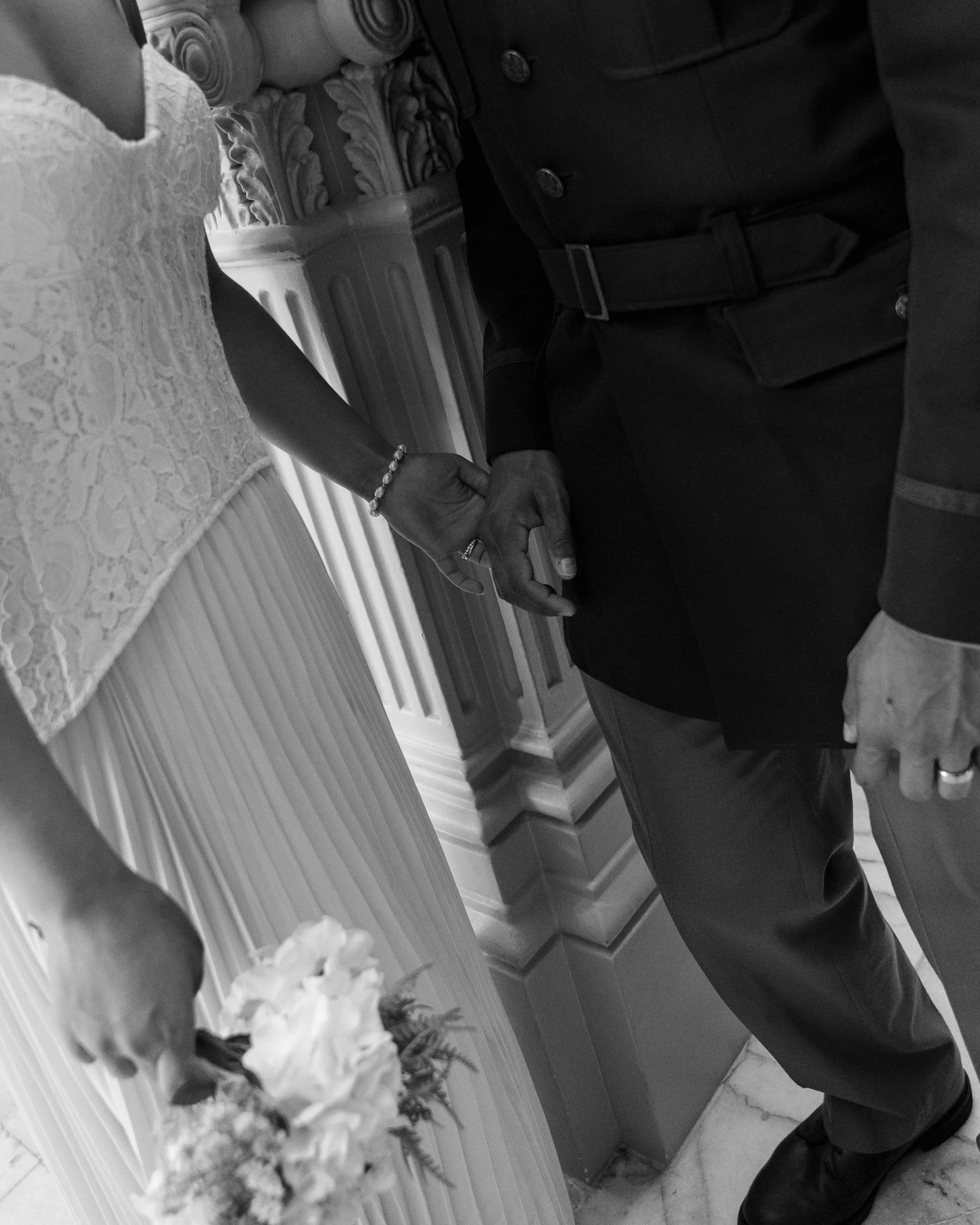 Close-up of a couple holding hands during their wedding, with the woman in a lace dress and jewelry and the man in a dark suit, in an ornate room.