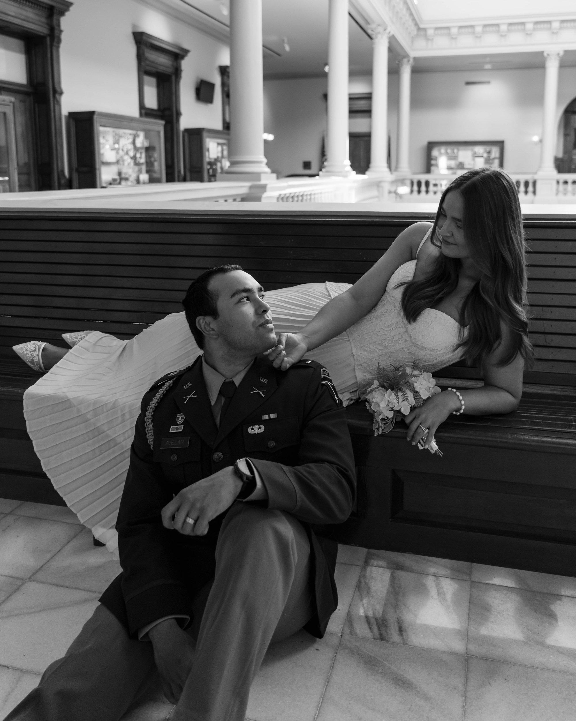 A man in military uniform sitting on the floor looking up at a woman in a wedding dress who is lying on a bench above him, in a large indoor space with columns and wooden paneling.