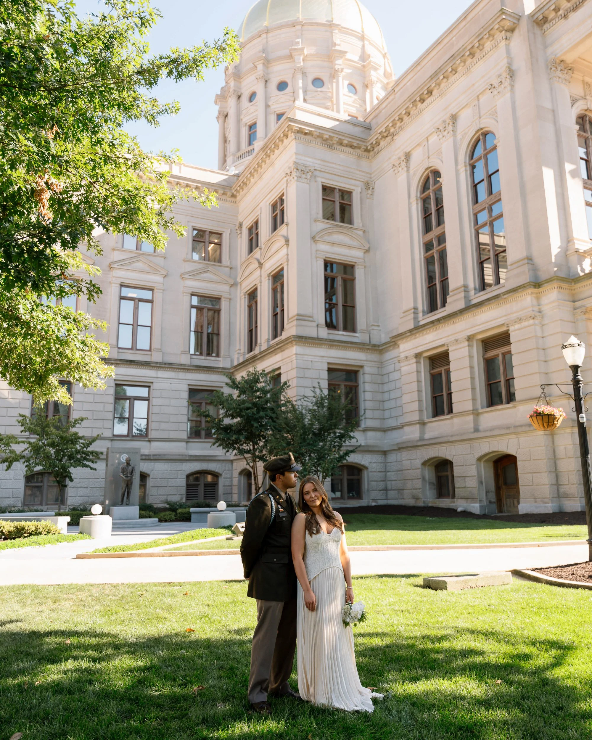 A newlywed couple standing on a green lawn in front of a historic government building with a domed roof, large windows, and ornate architecture. The bride is holding a bouquet of white flowers, and the groom is in a military uniform.