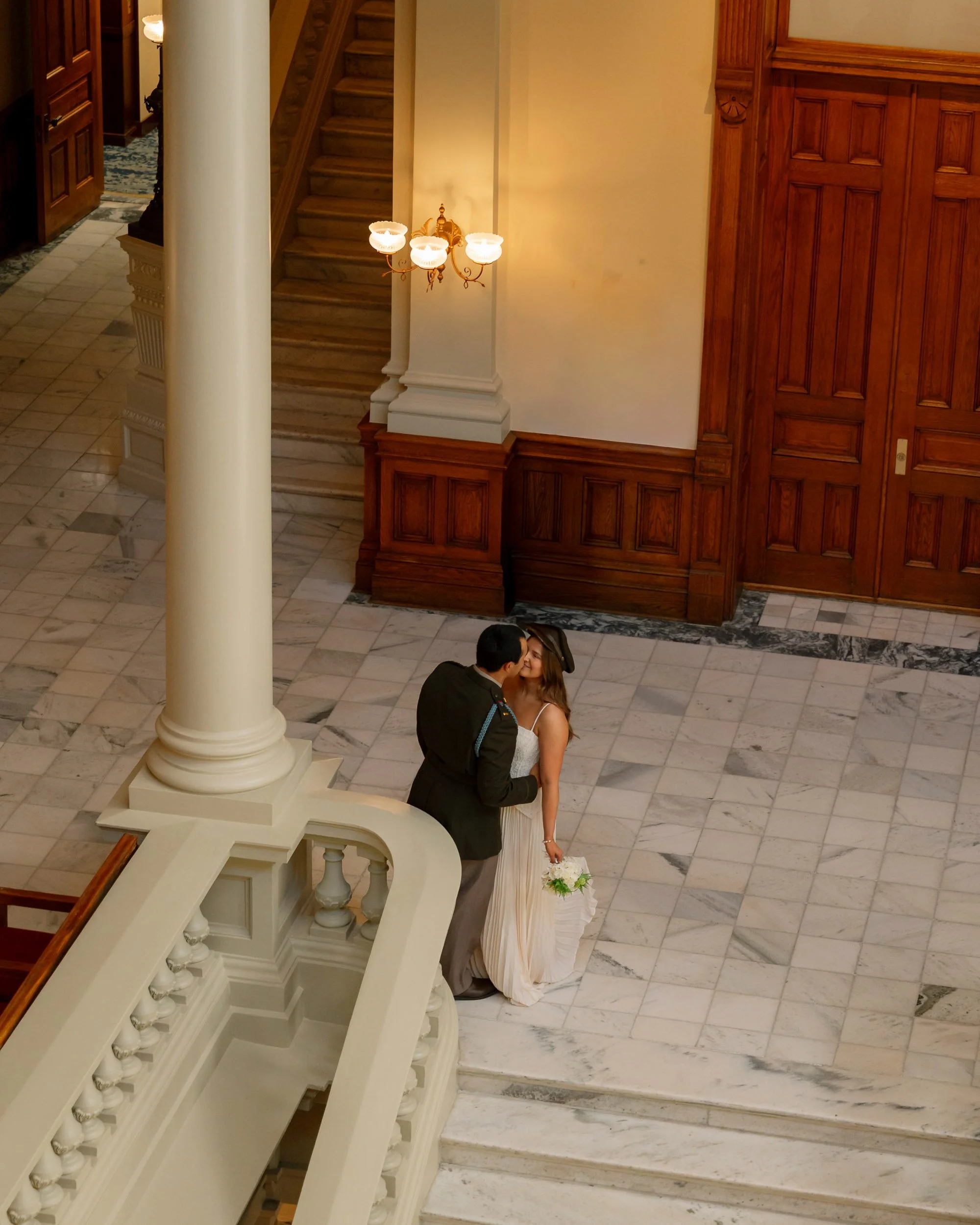 A bride and groom share a kiss in a grand historic building with marble floors, wooden paneling, and a large staircase.