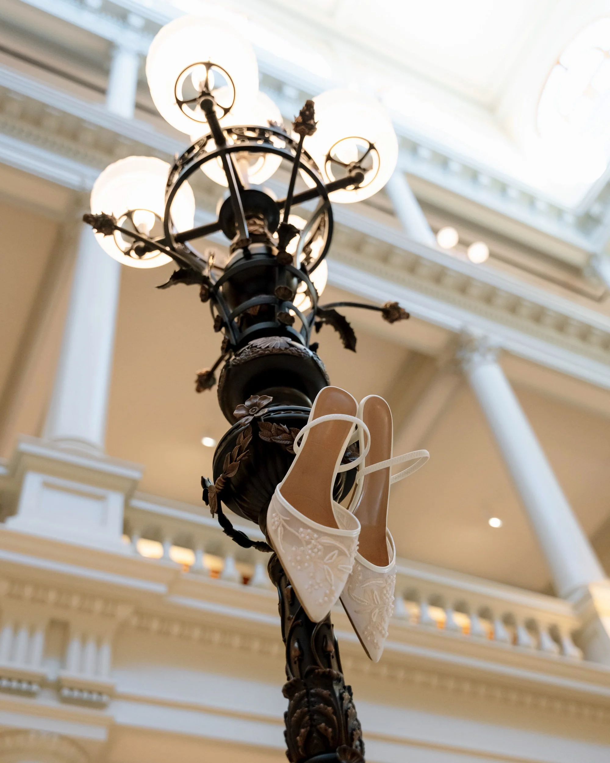A decorative street lamp with white high-heeled shoes hanging from it, inside a building with classical architecture and a glass ceiling.