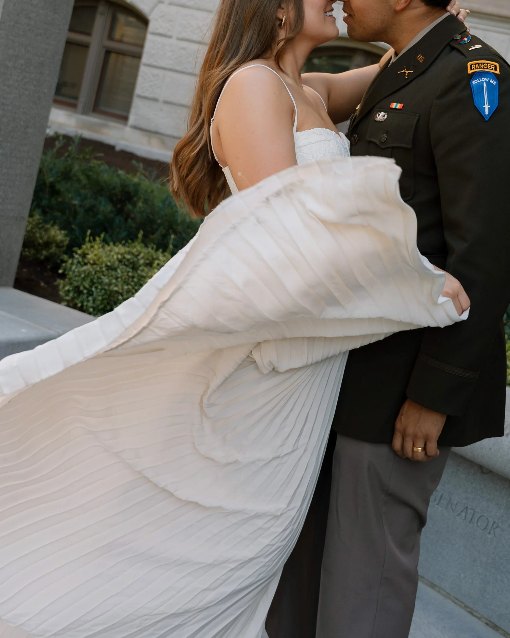 A newlywed couple embracing each other during their wedding, with the bride in a white dress and the groom in a military uniform, standing outside in front of a stone building.