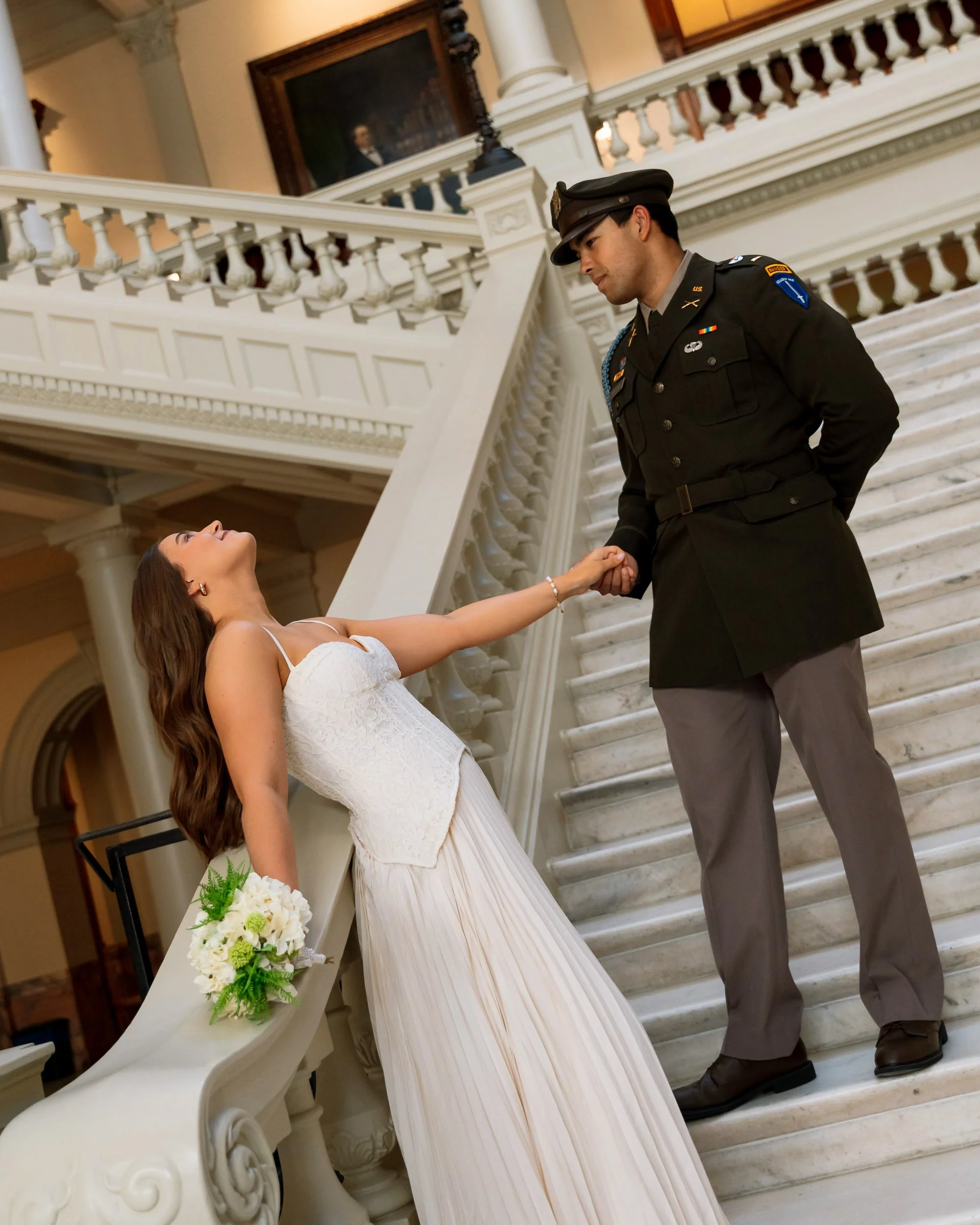 A woman in a white wedding dress leaning on a staircase railing, holding a bouquet, as she holds hands with a man in a military uniform on the staircase.