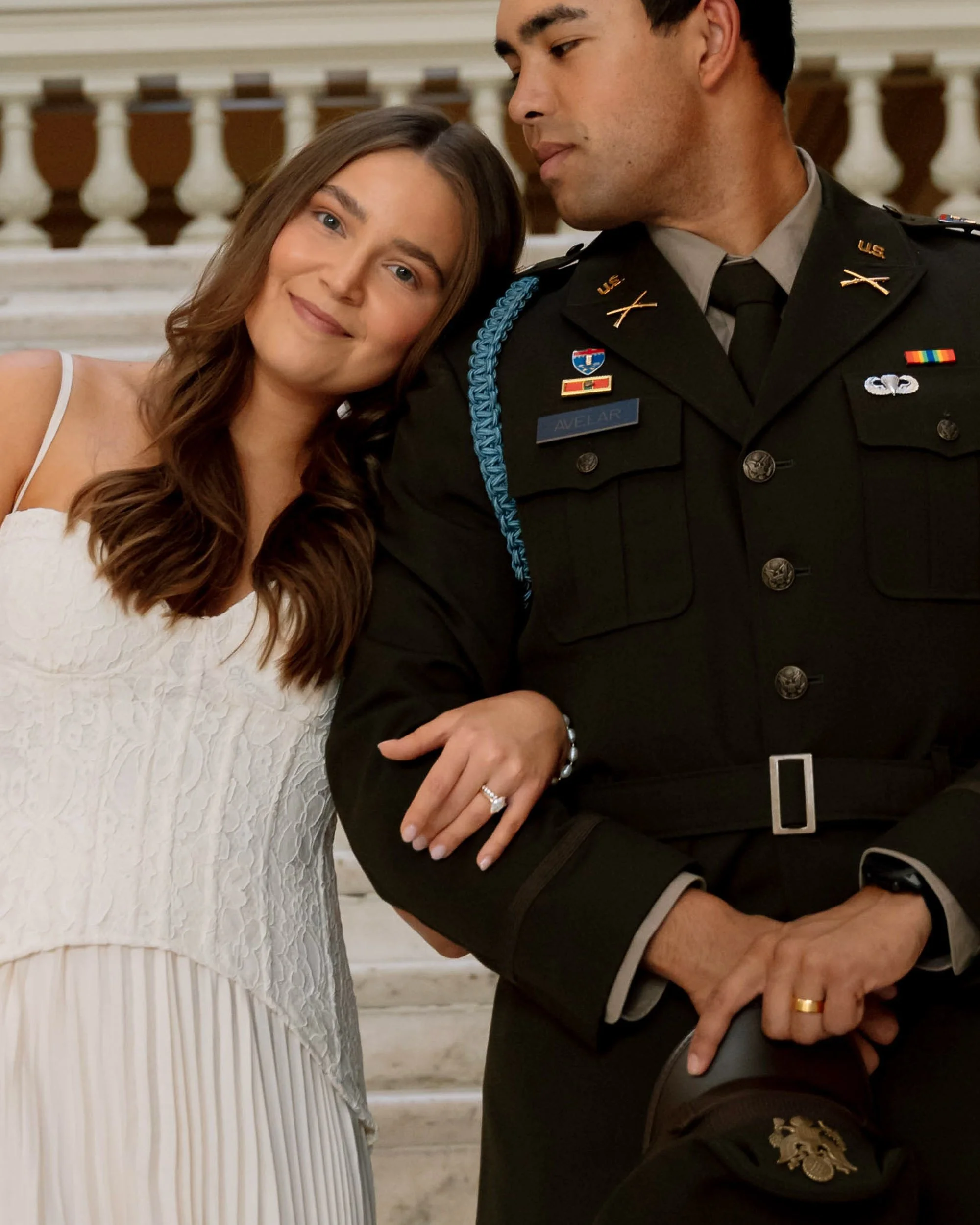 A woman in a white dress with brown hair standing close to a man in a U.S. military uniform on a staircase.