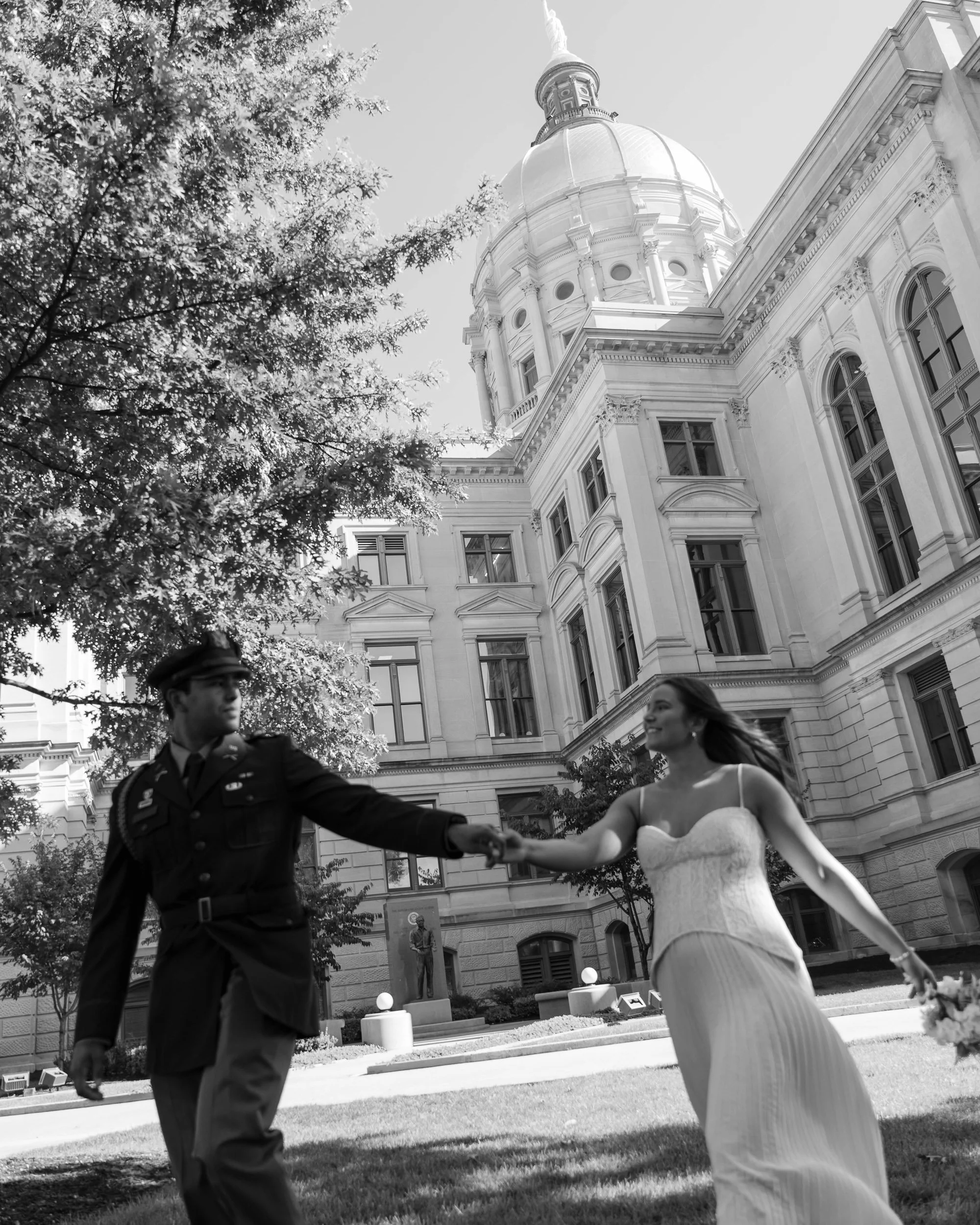A couple in wedding attire holding hands on a lawn in front of a large, historic government building with a dome and tall windows.