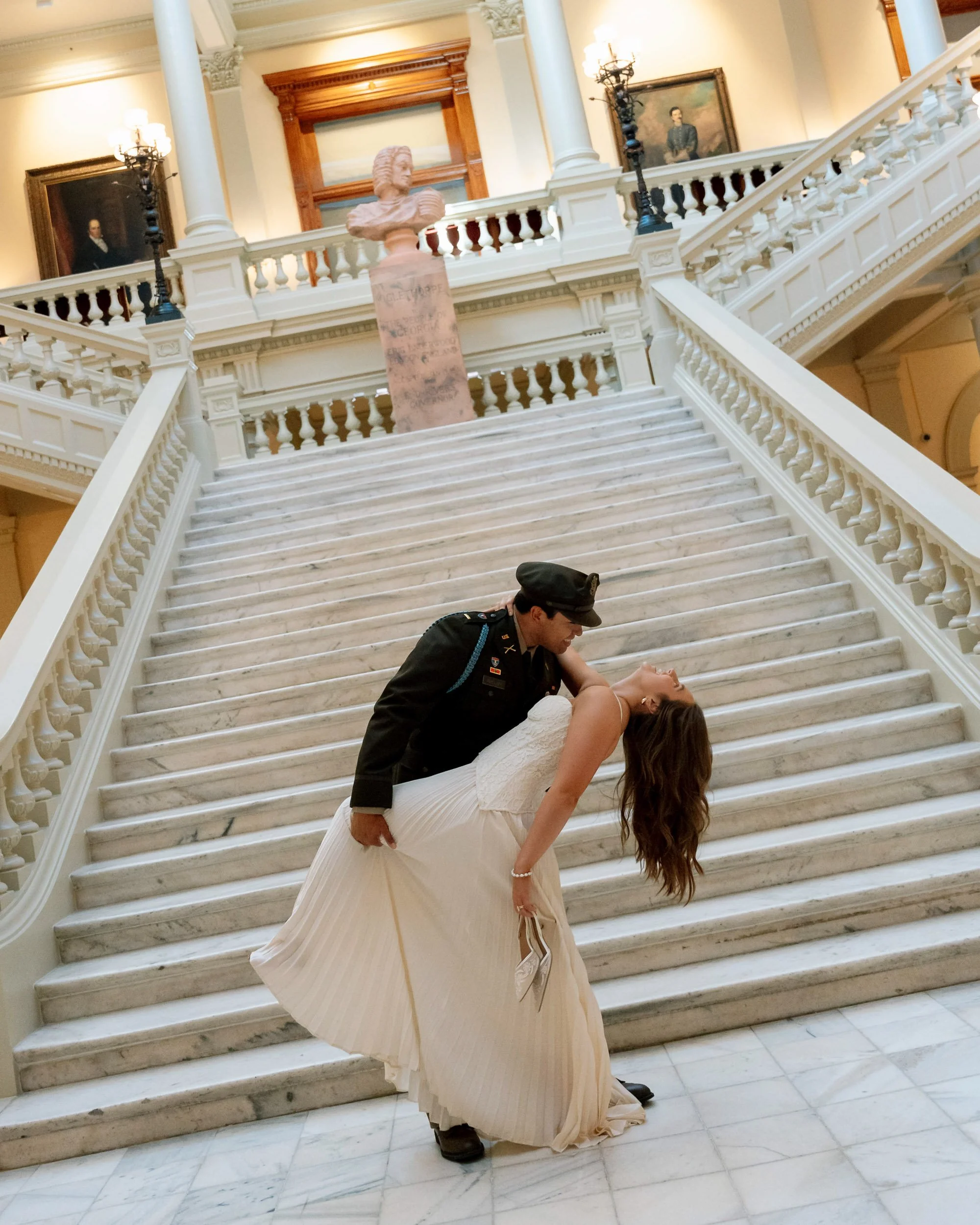 A man dressed as a soldier in uniform dips a woman in a white dress as part of a dance on a grand marble staircase inside a building with classical decor, framed portraits, and bust sculptures.