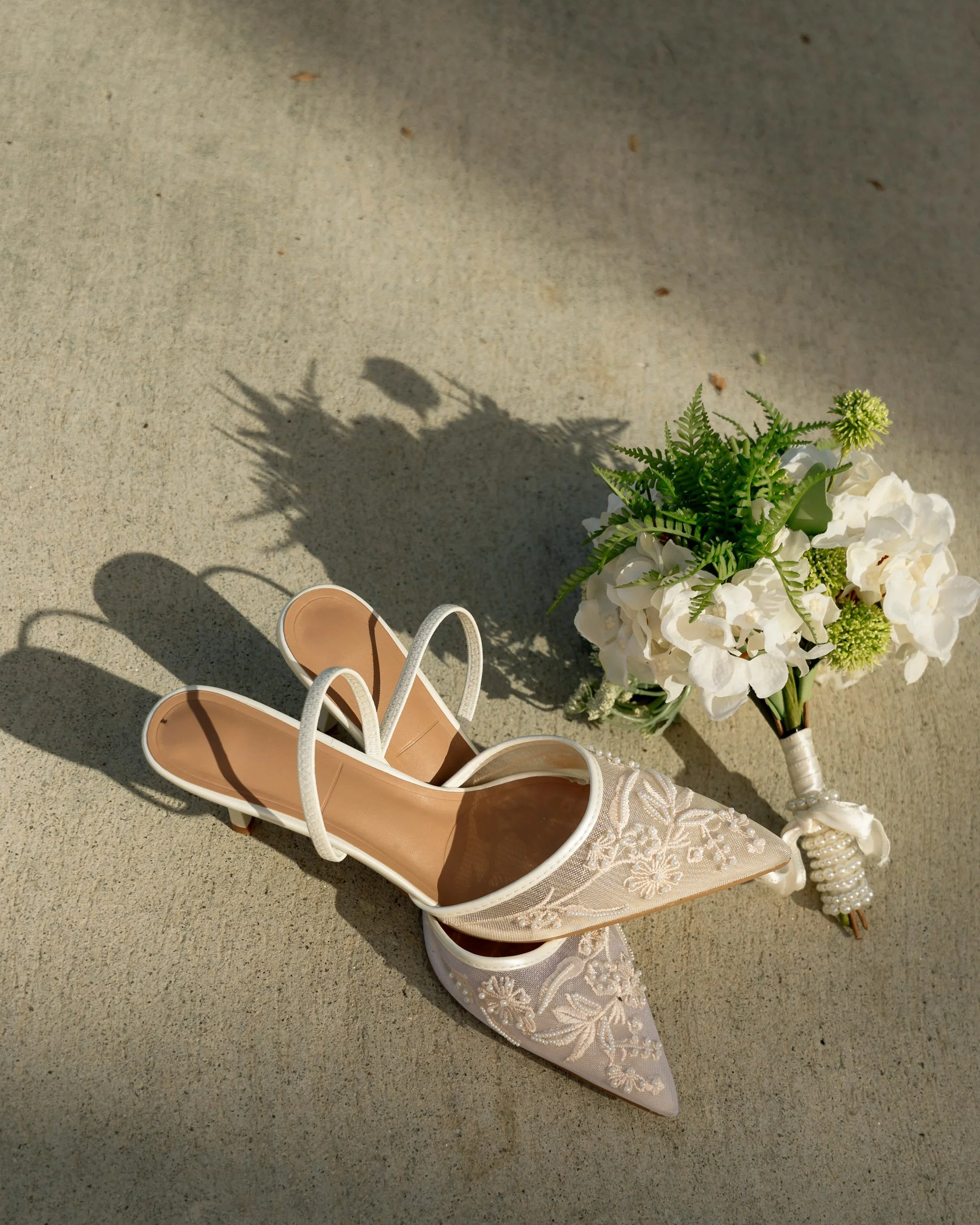 A pair of beige high-heeled shoes with lace embroidery and a floral bouquet with white flowers and green foliage on a concrete surface.