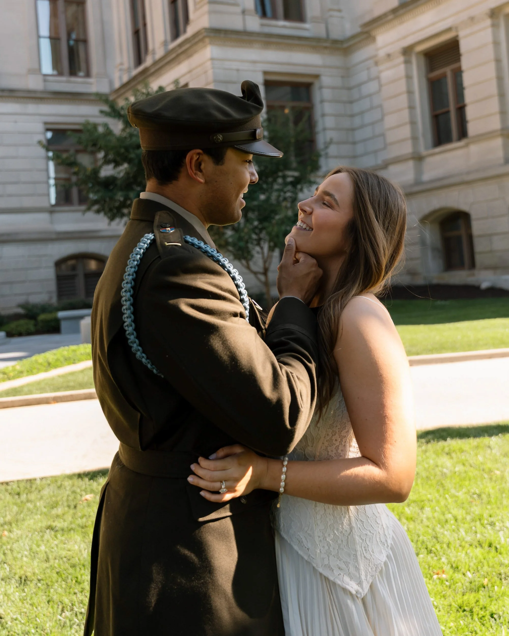 A military officer and a woman in a wedding dress sharing a romantic moment outdoors, with the officer holding the woman's chin and smiling.