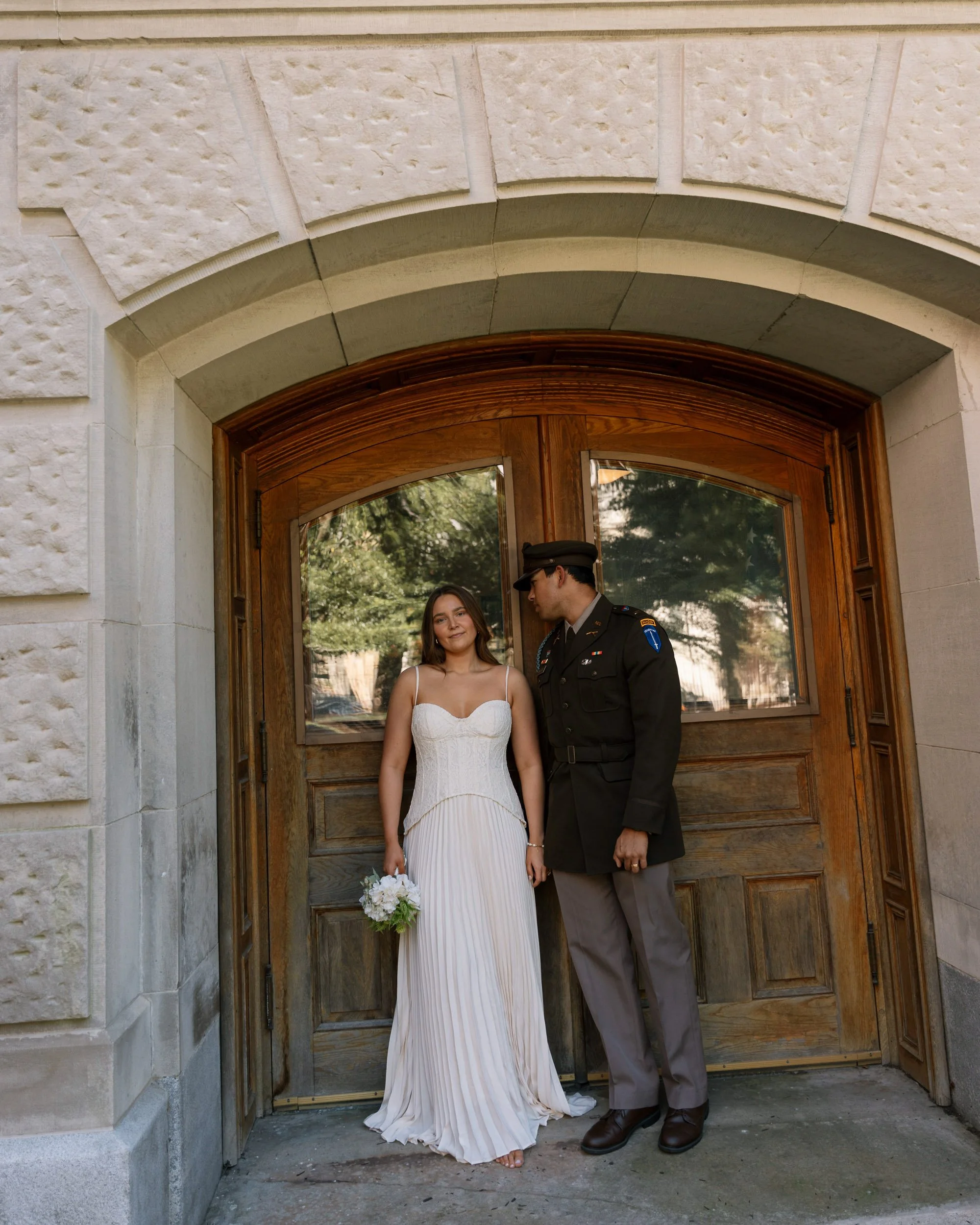 A woman in a white dress holding a bouquet standing next to a man in a military uniform in front of a wooden door with stone framing, reflecting trees in the glass.