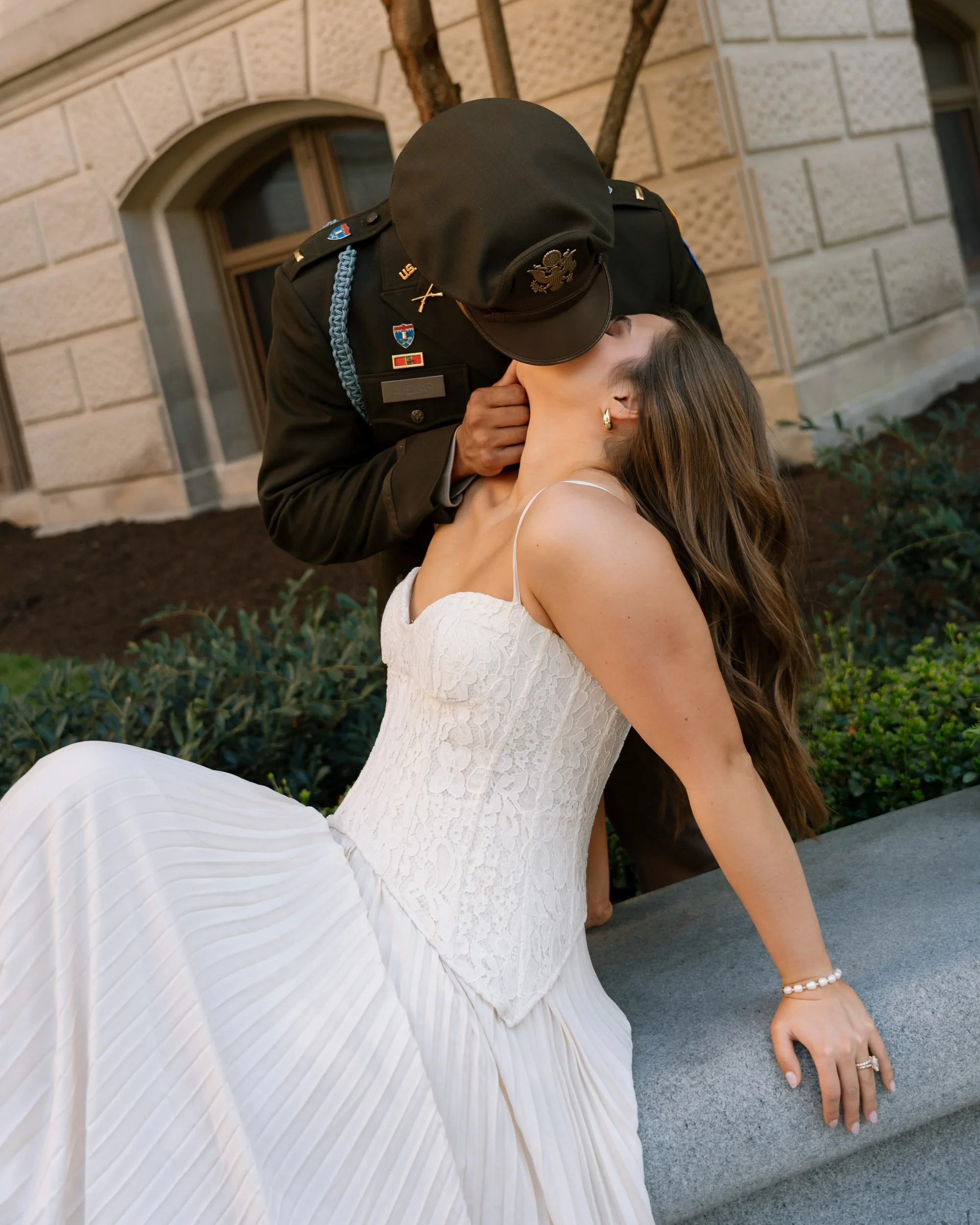 A soldier in uniform leaning down to kiss a woman in a white dress, sitting on a concrete bench outdoors near a building with arched windows.